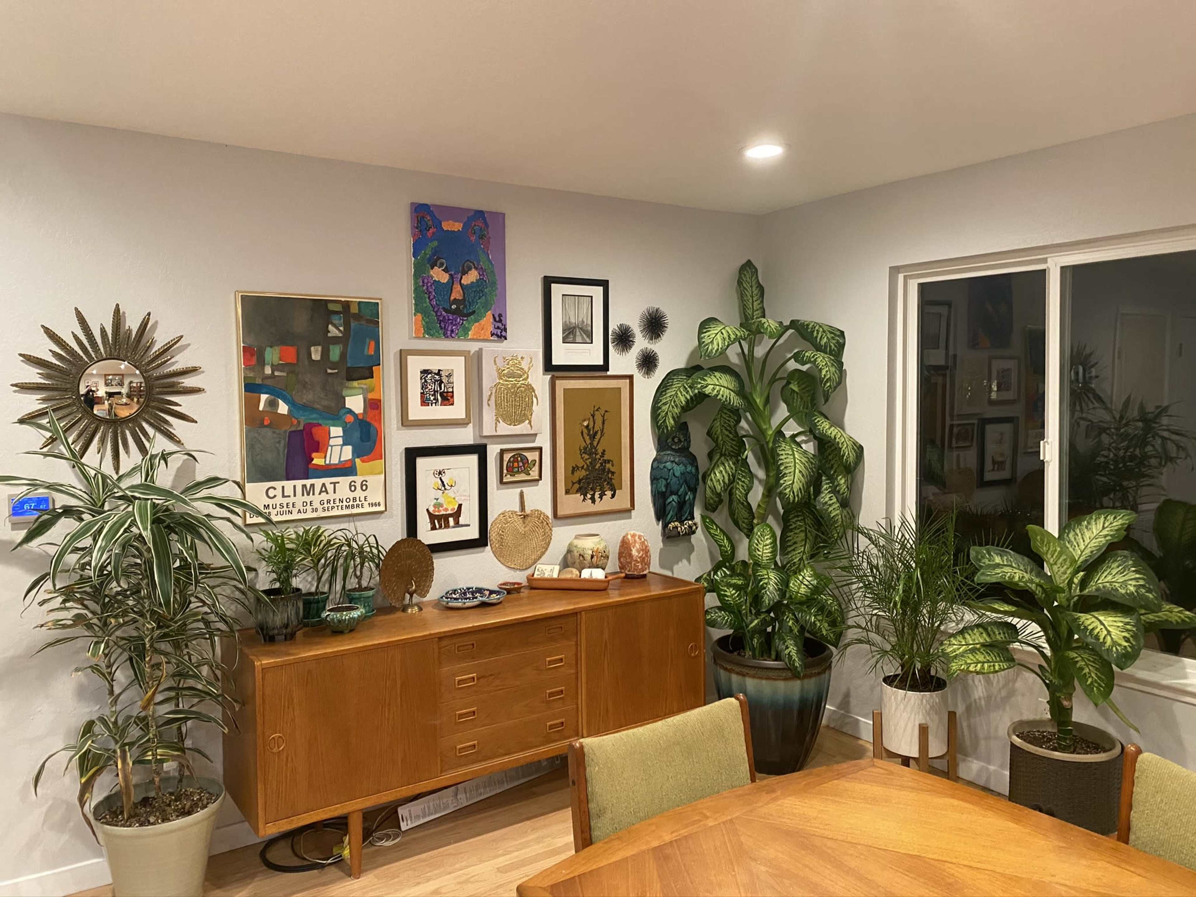 A well-decorated living space with a wooden sideboard displaying various decorative objects and surrounded by plants, while a gallery wall of framed artworks is visible behind it.