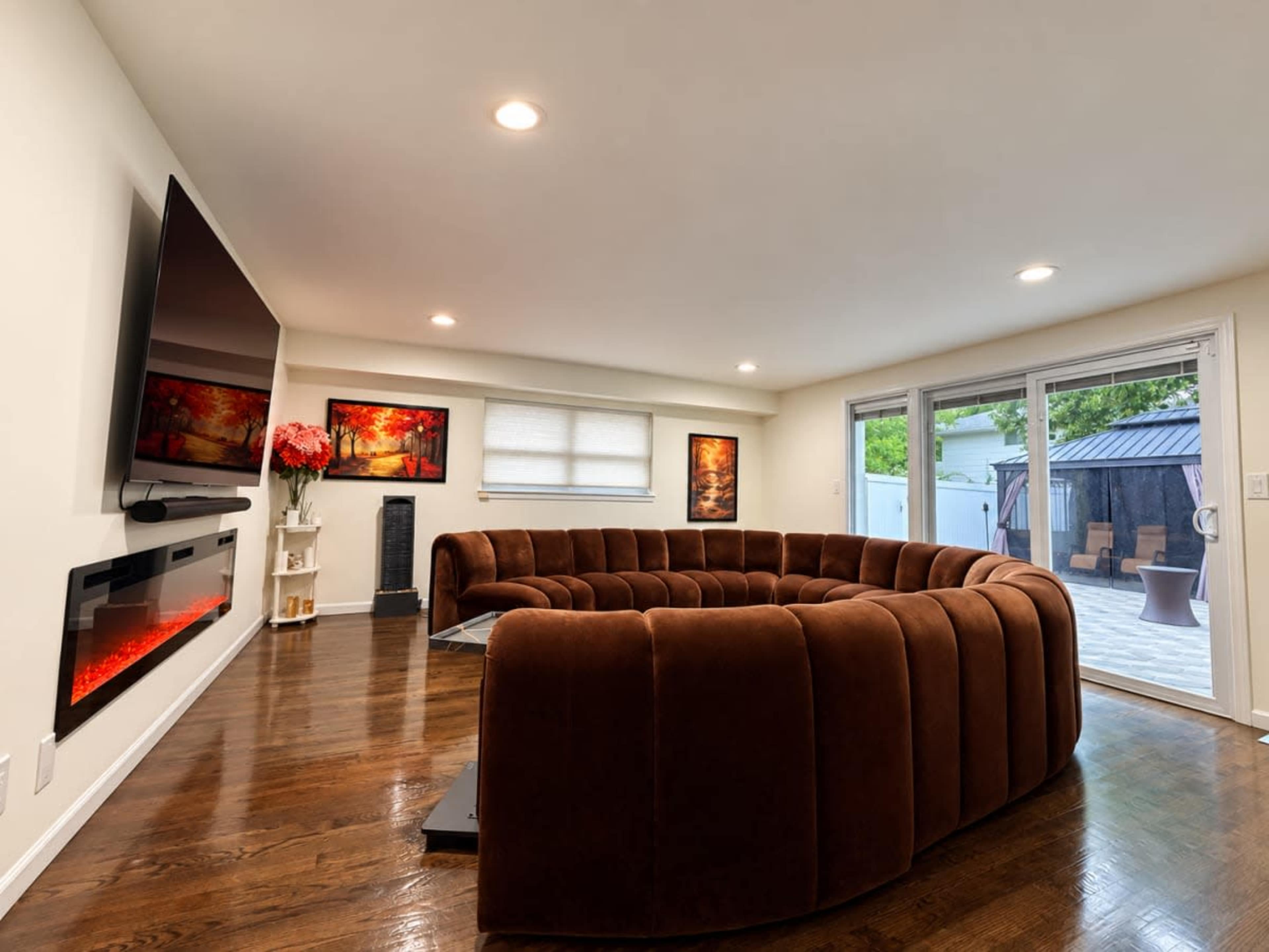 The image shows a modern living room with a curved brown sofa, a large wall-mounted TV, an electric fireplace, and sliding glass doors leading to an outdoor area.