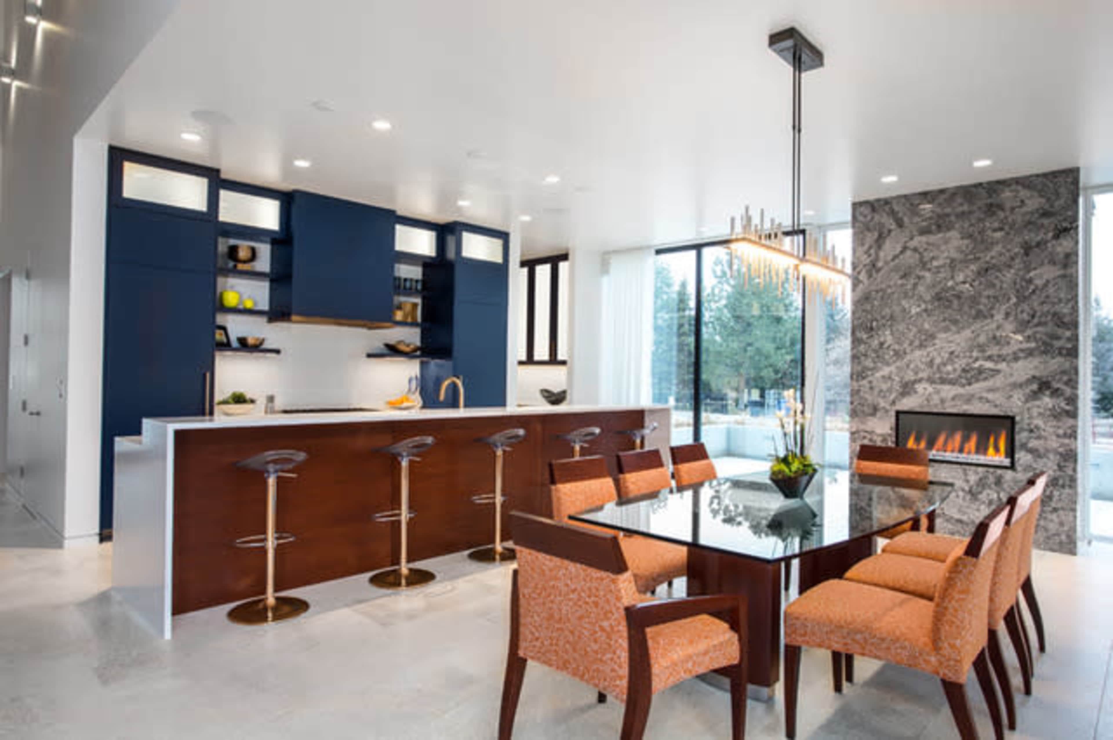 The image shows a modern kitchen and dining area featuring a large table surrounded by orange upholstered chairs, a marble fireplace, and a kitchen island with barstools.
