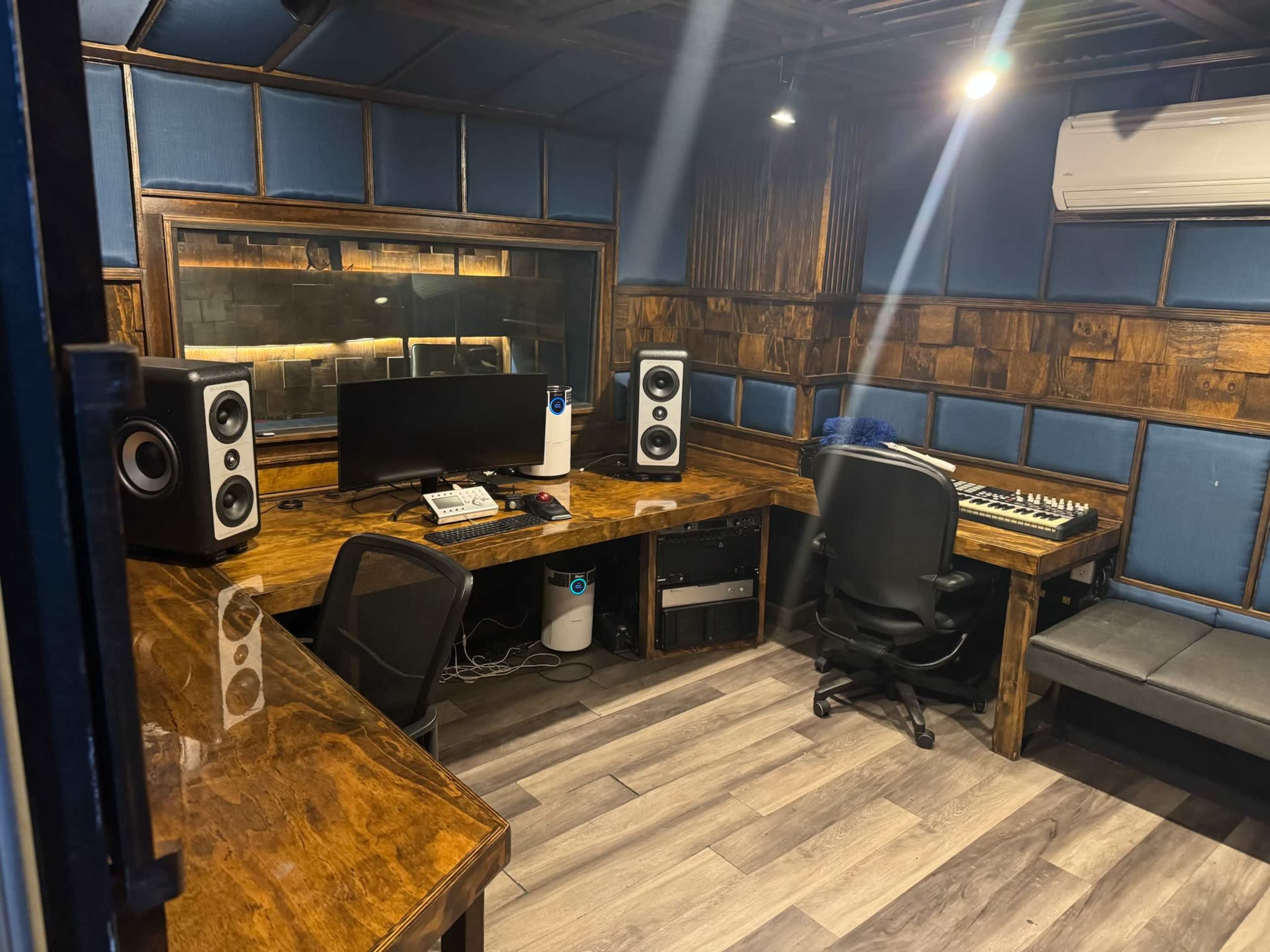 The image shows a well-equipped recording studio featuring a wooden desk, computer monitors, studio speakers, and soundproof walls.