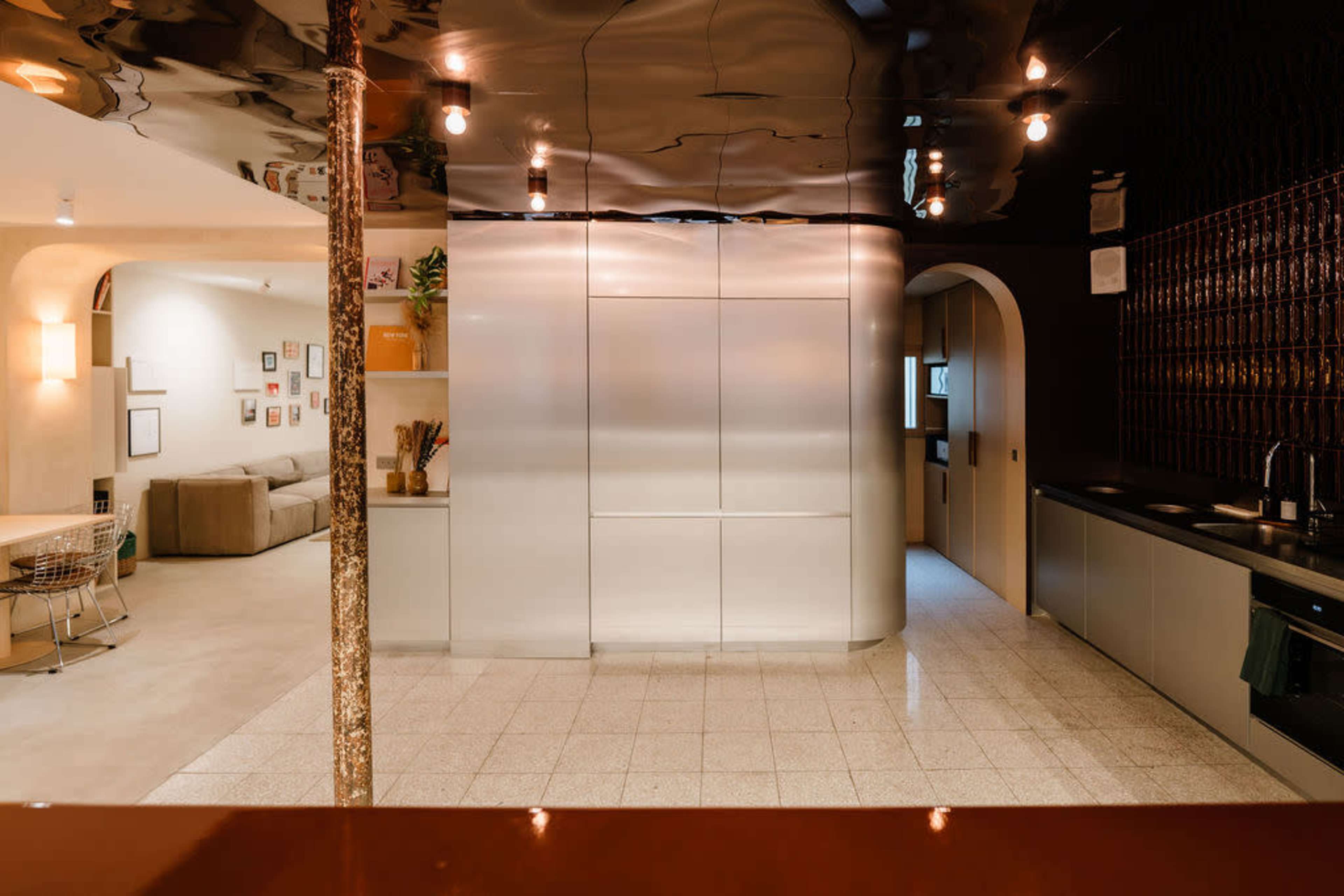 The image shows a modern kitchen with sleek metallic cabinets, a light-colored tile floor, and a spacious layout that connects to a living area.