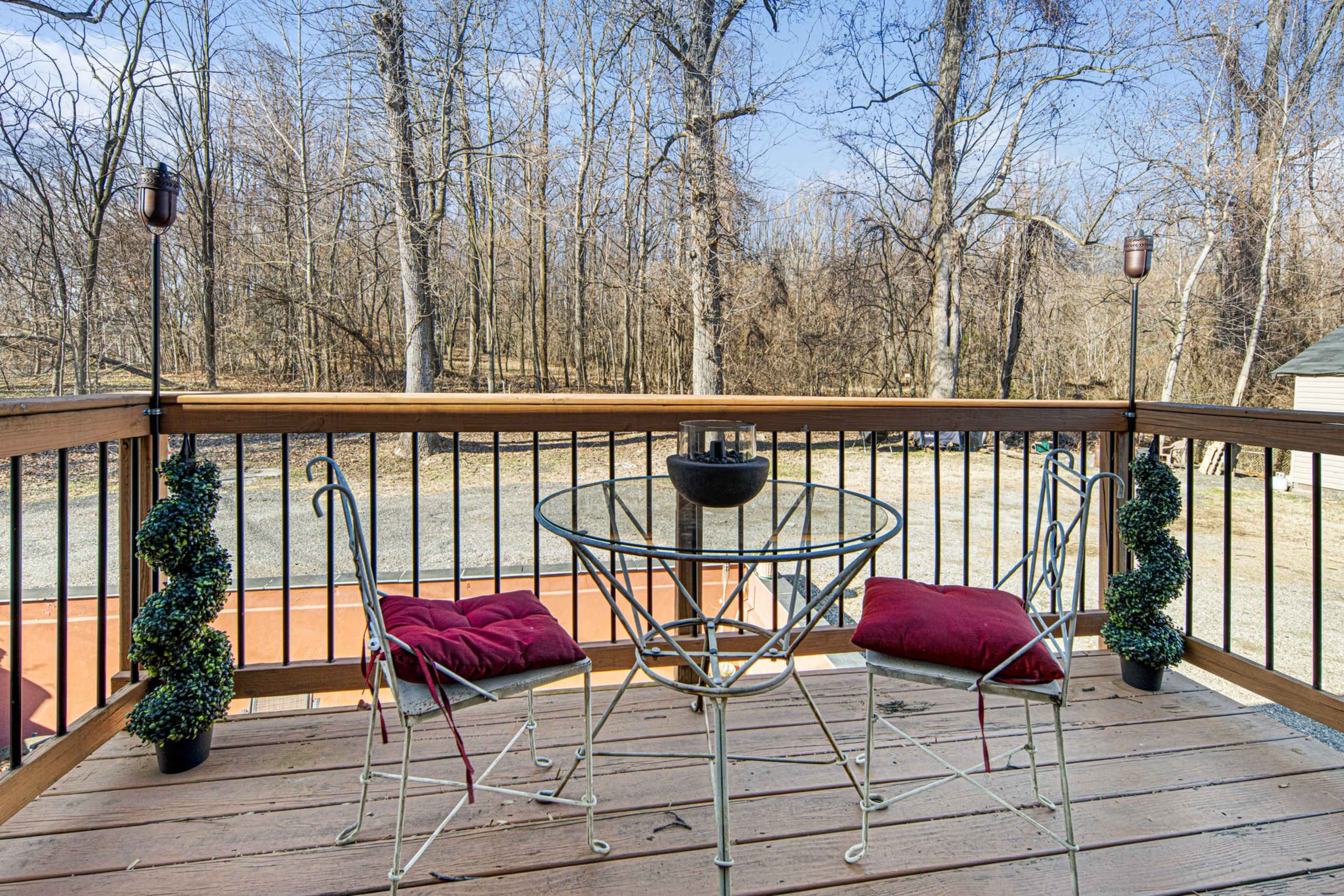 A small outdoor deck features a round table and two metal chairs with red cushions, surrounded by potted greenery and a view of bare trees in the background.
