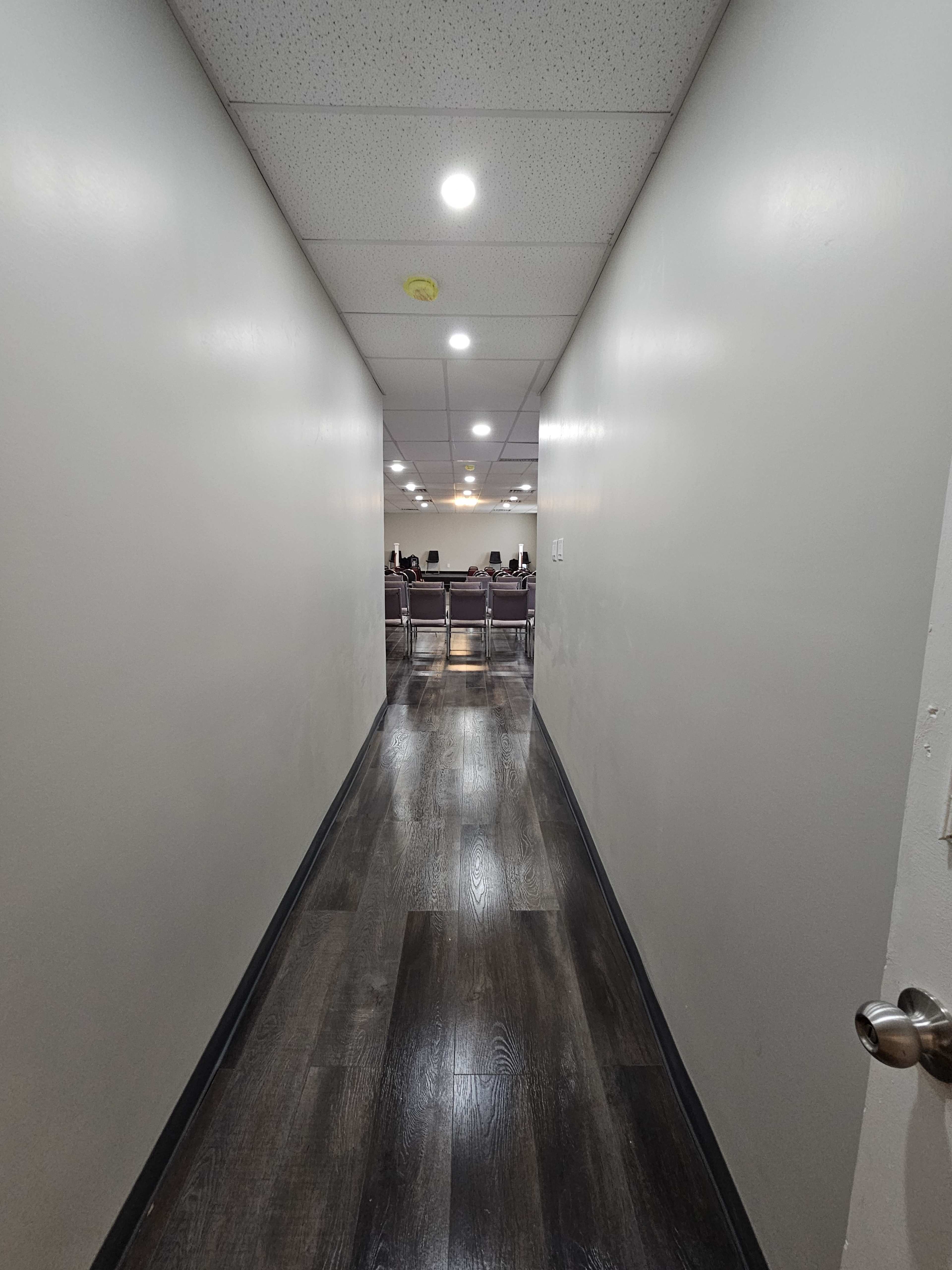 The image shows a narrow hallway leading to a room with several chairs lined up in the background.