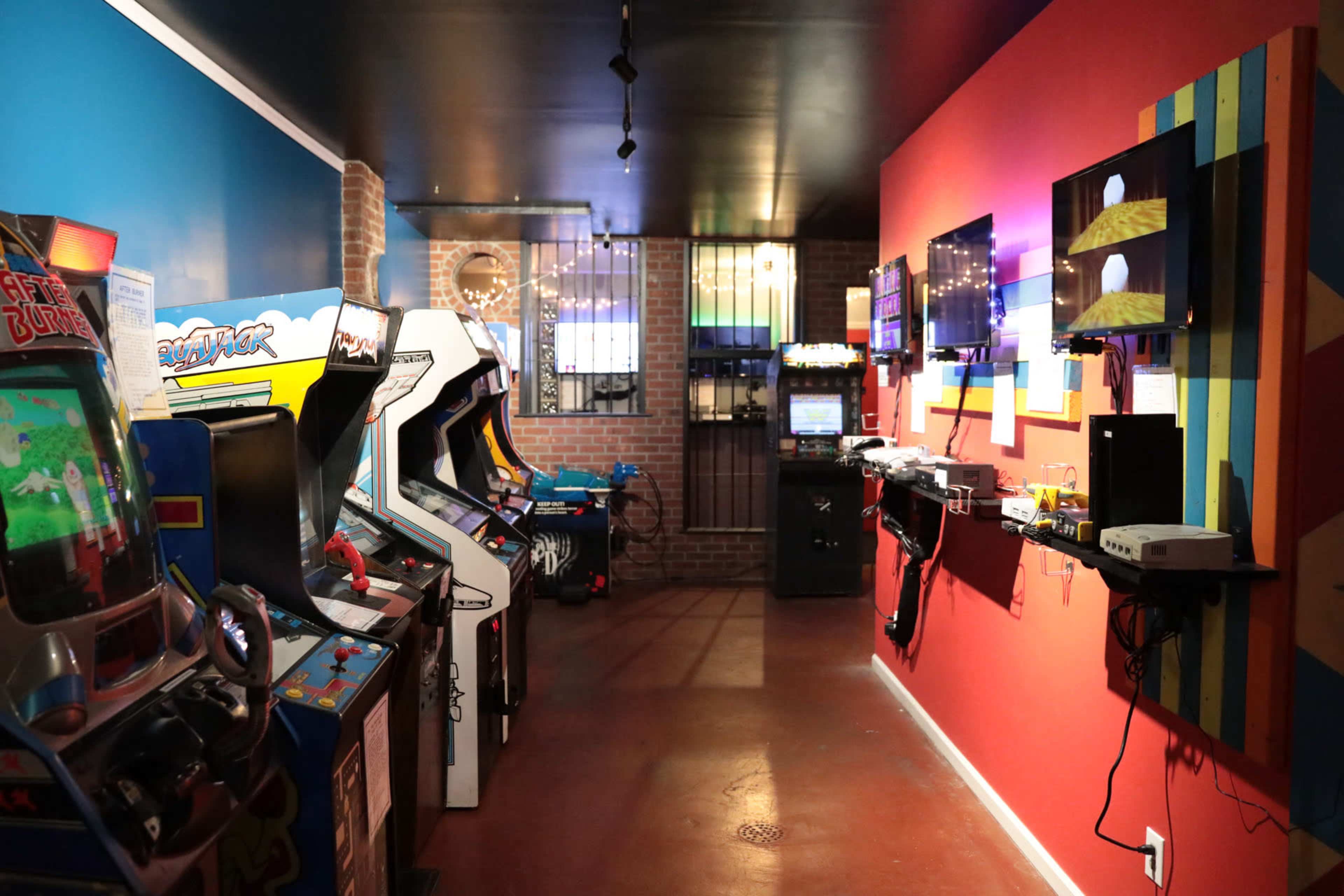 A retro arcade room with a row of vintage arcade machines on one side and wall-mounted gaming consoles on the opposite side.