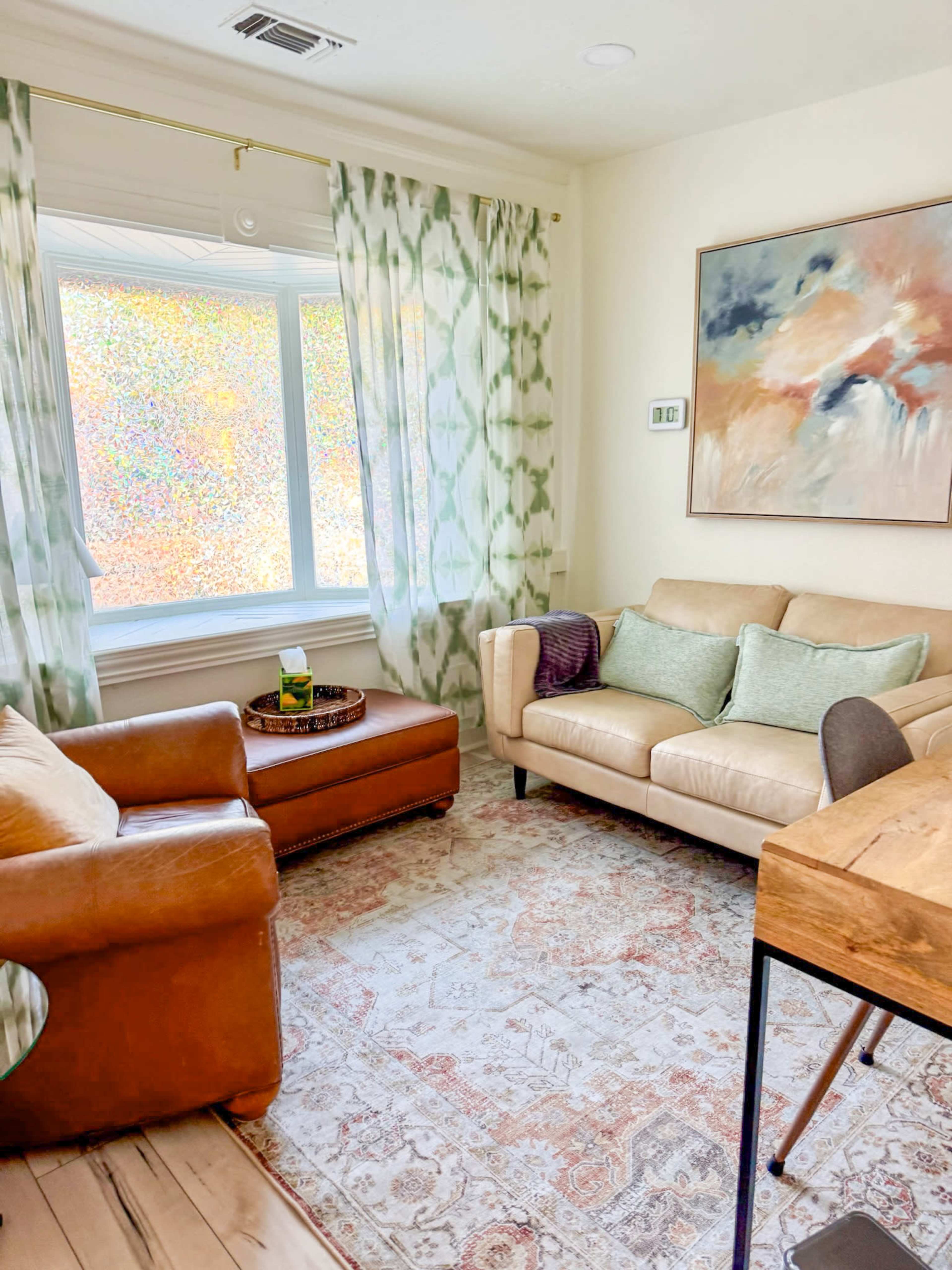 A cozy living room features a light brown leather sofa, a wooden coffee table, and a large window with sheer green curtains letting in colorful natural light.