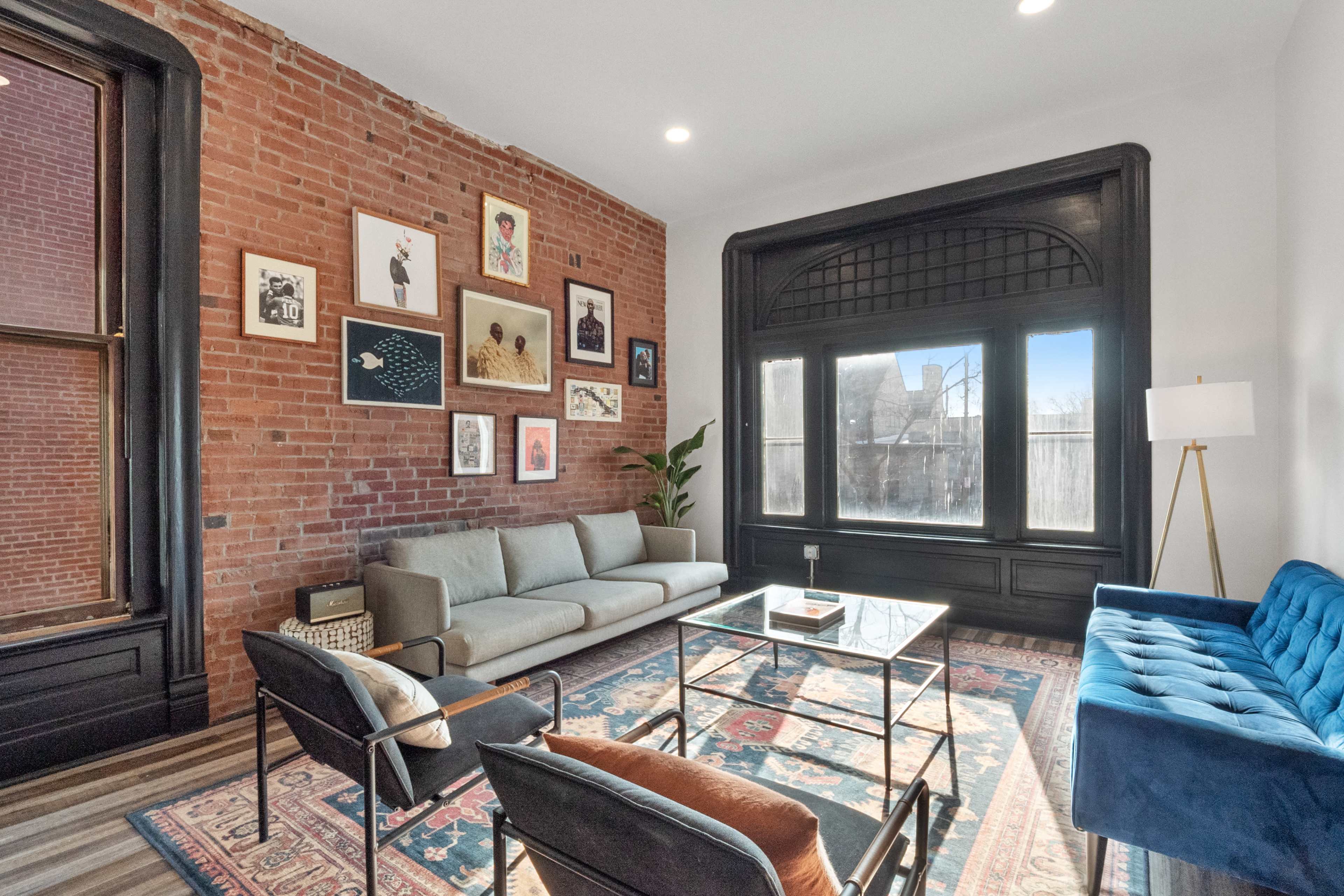A modern living room with exposed brick walls, a green sofa, a blue couch, a glass coffee table, and a large window alongside framed artwork.