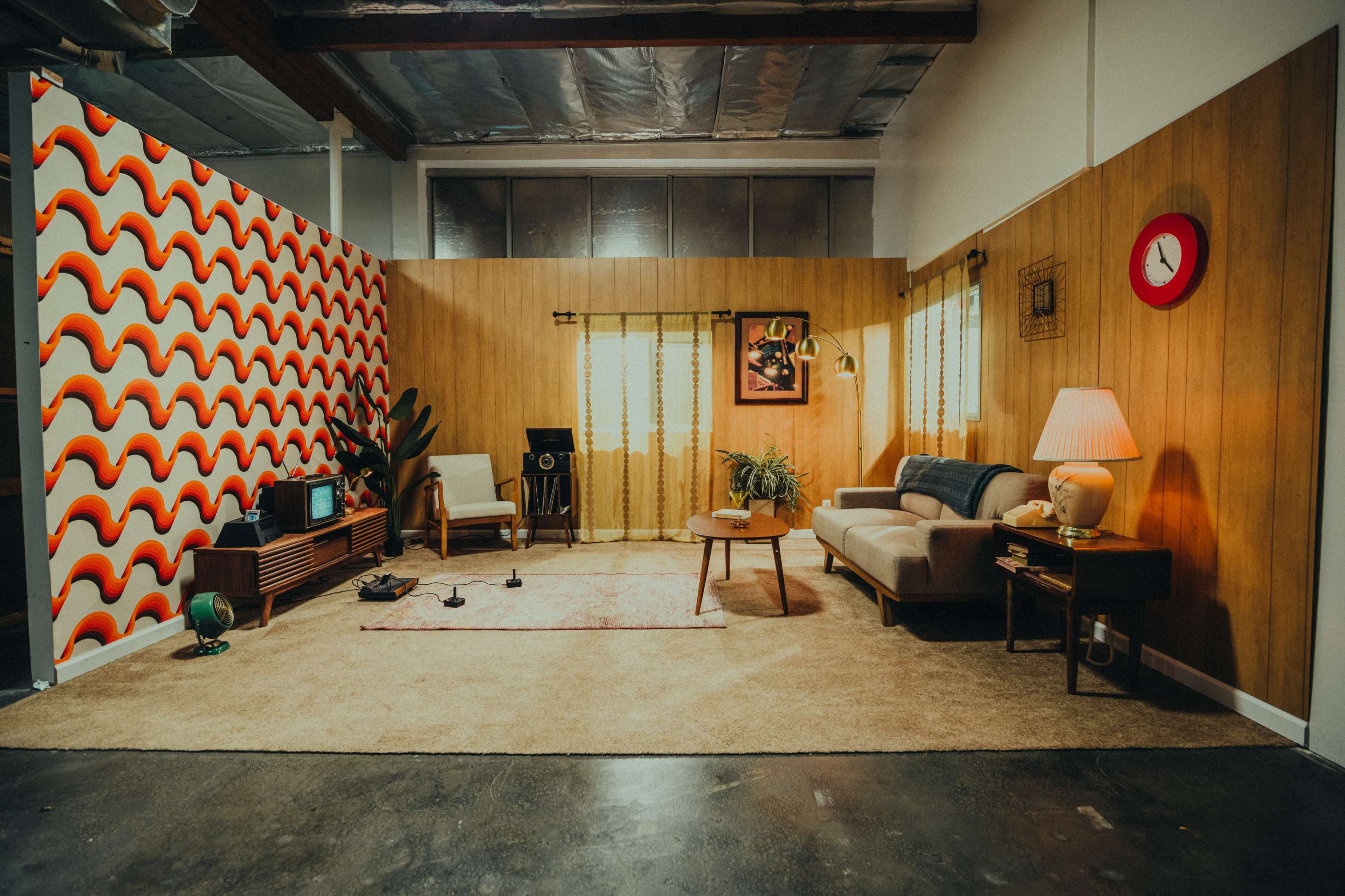 A retro-styled living room with a patterned wallpaper, beige carpeting, and a mix of vintage furniture and decor items.