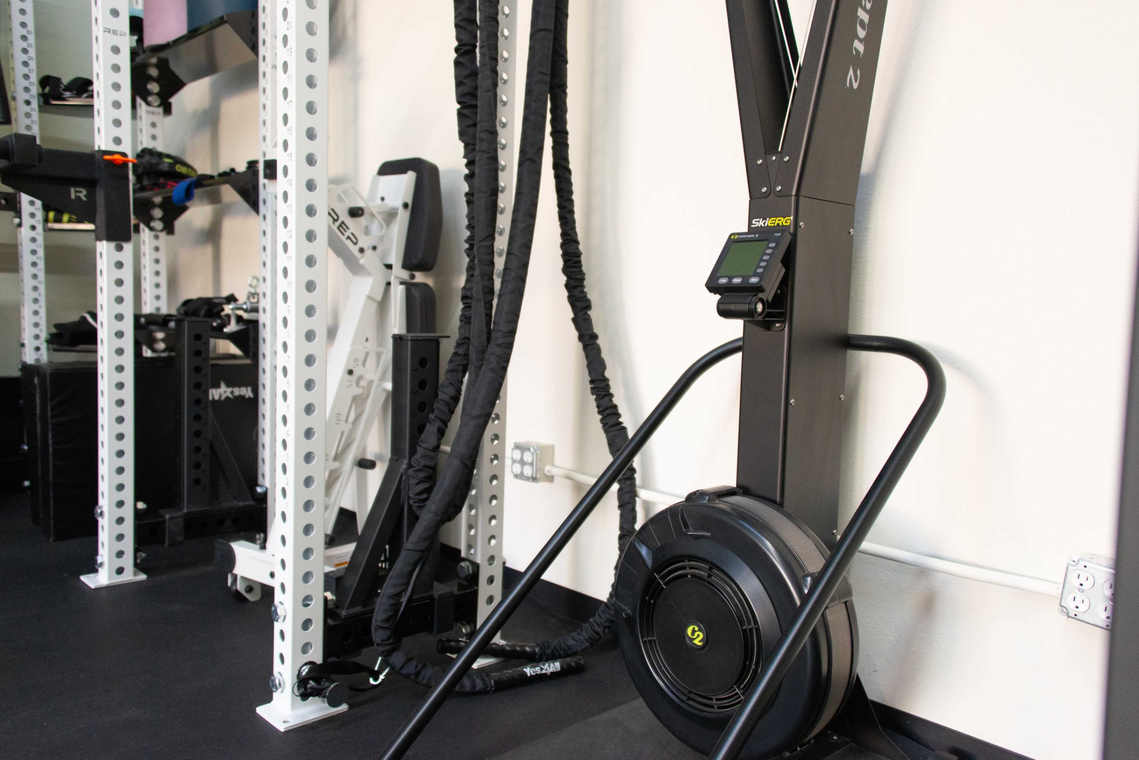 The image shows a gym space featuring a rowing machine, various workout equipment, and exercise cables mounted on the wall.