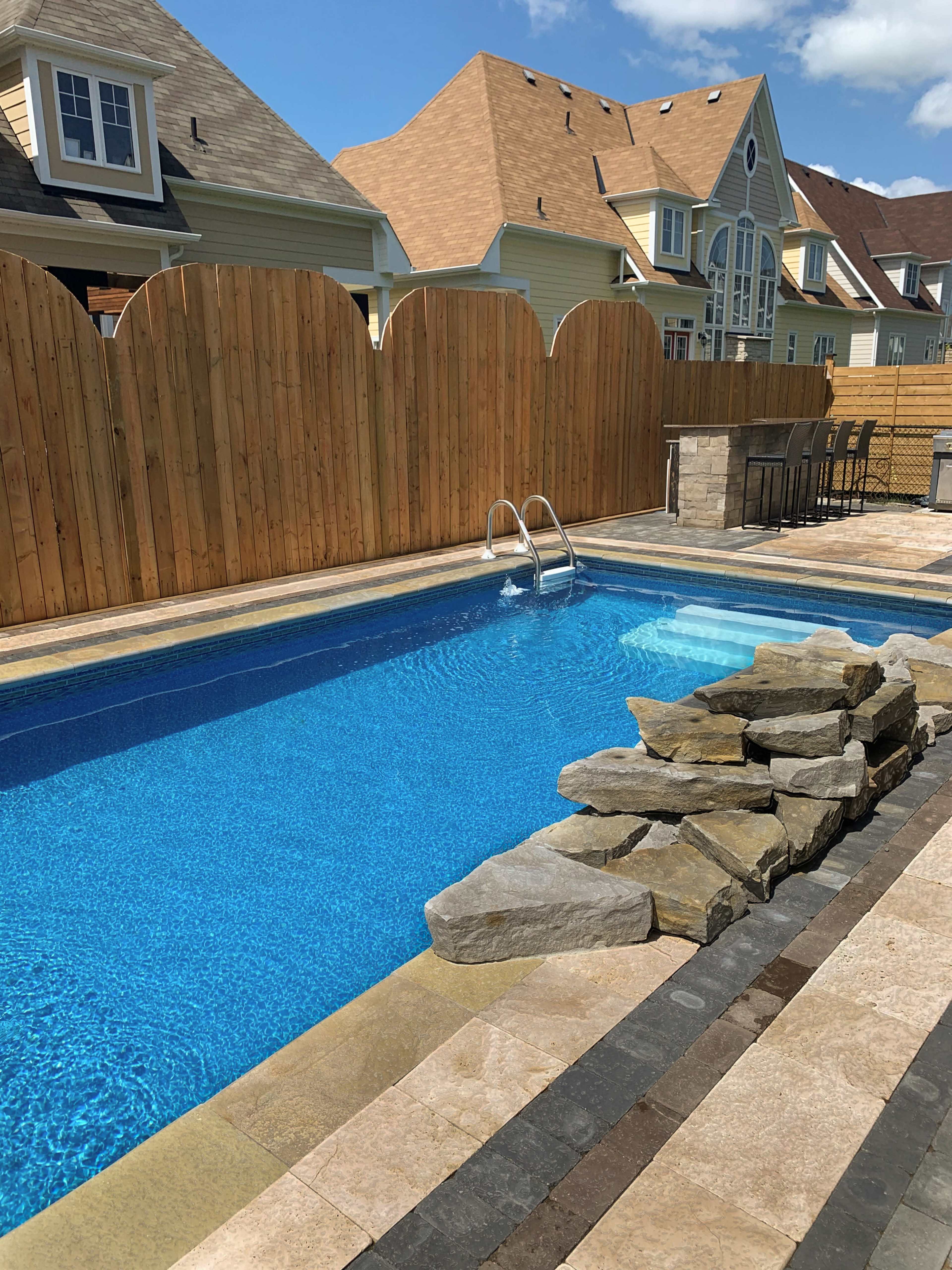 A rectangular swimming pool with a rock feature, surrounded by a stone patio and fenced by wooden panels, with houses visible in the background.