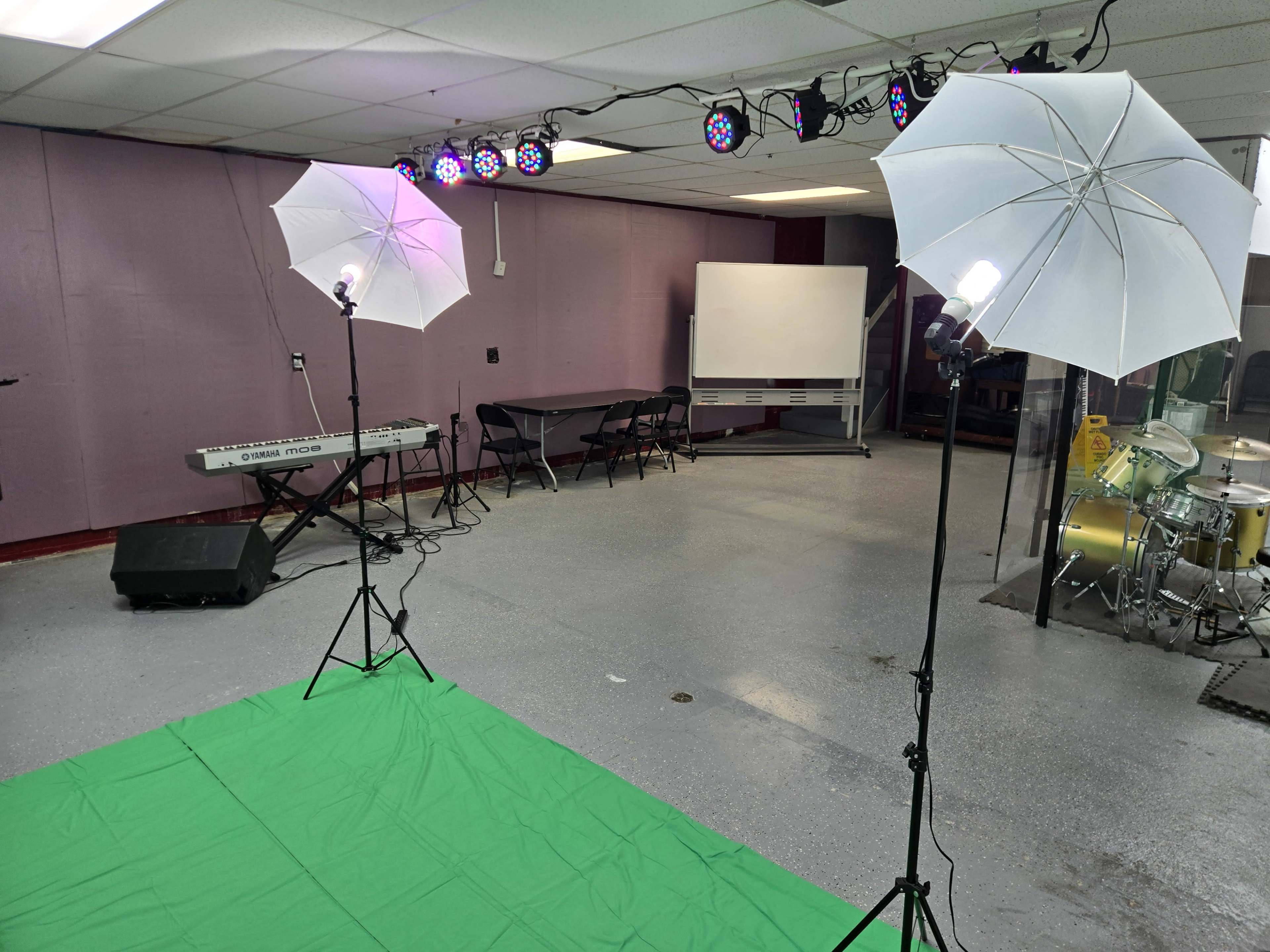The image shows a room setup for a performance or recording, featuring two softbox lights, a green backdrop, a keyboard, a whiteboard, and a drum set.