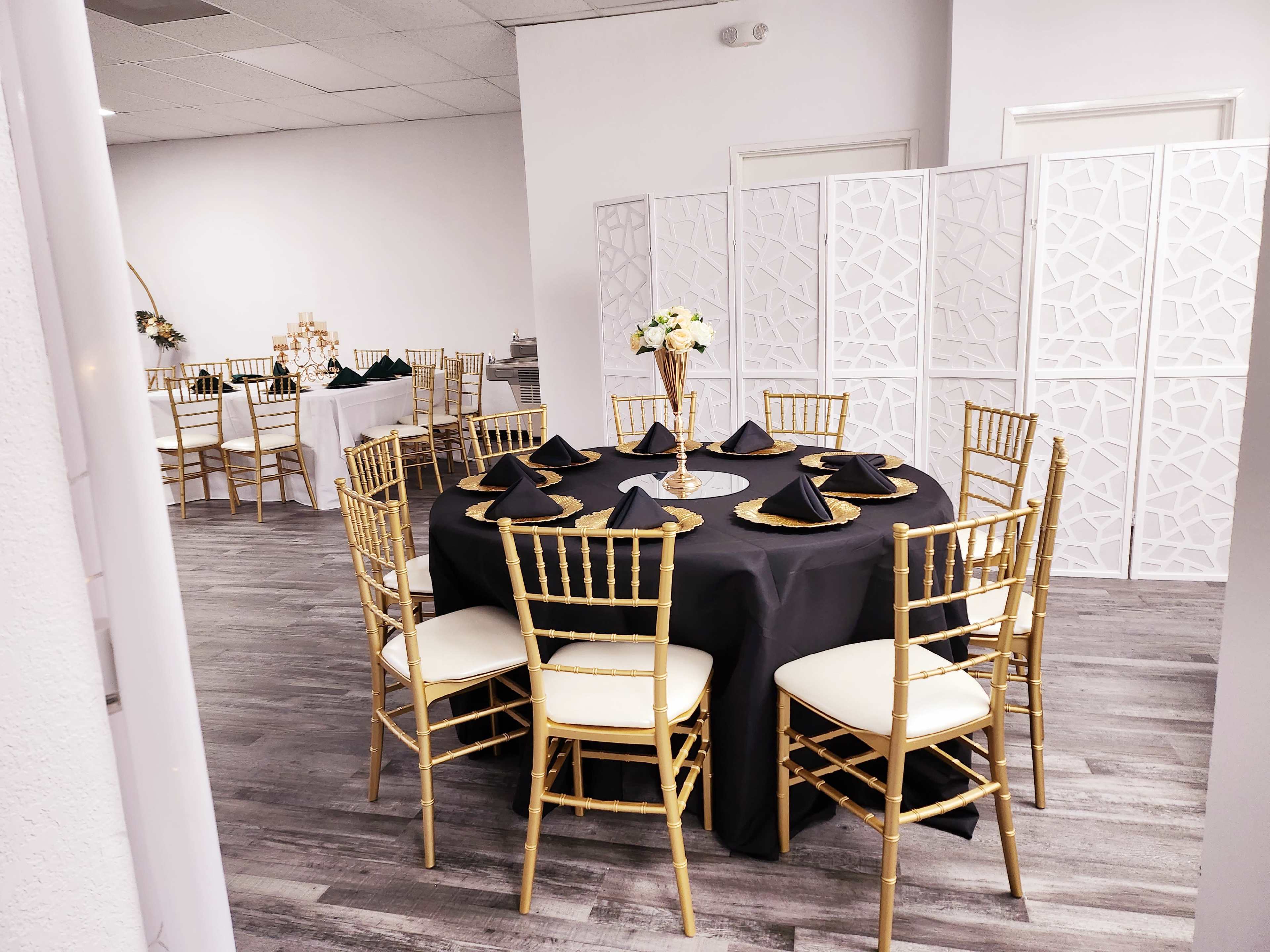 A round table set with black napkins and gold accents is positioned in a spacious event room featuring additional tables in the background.