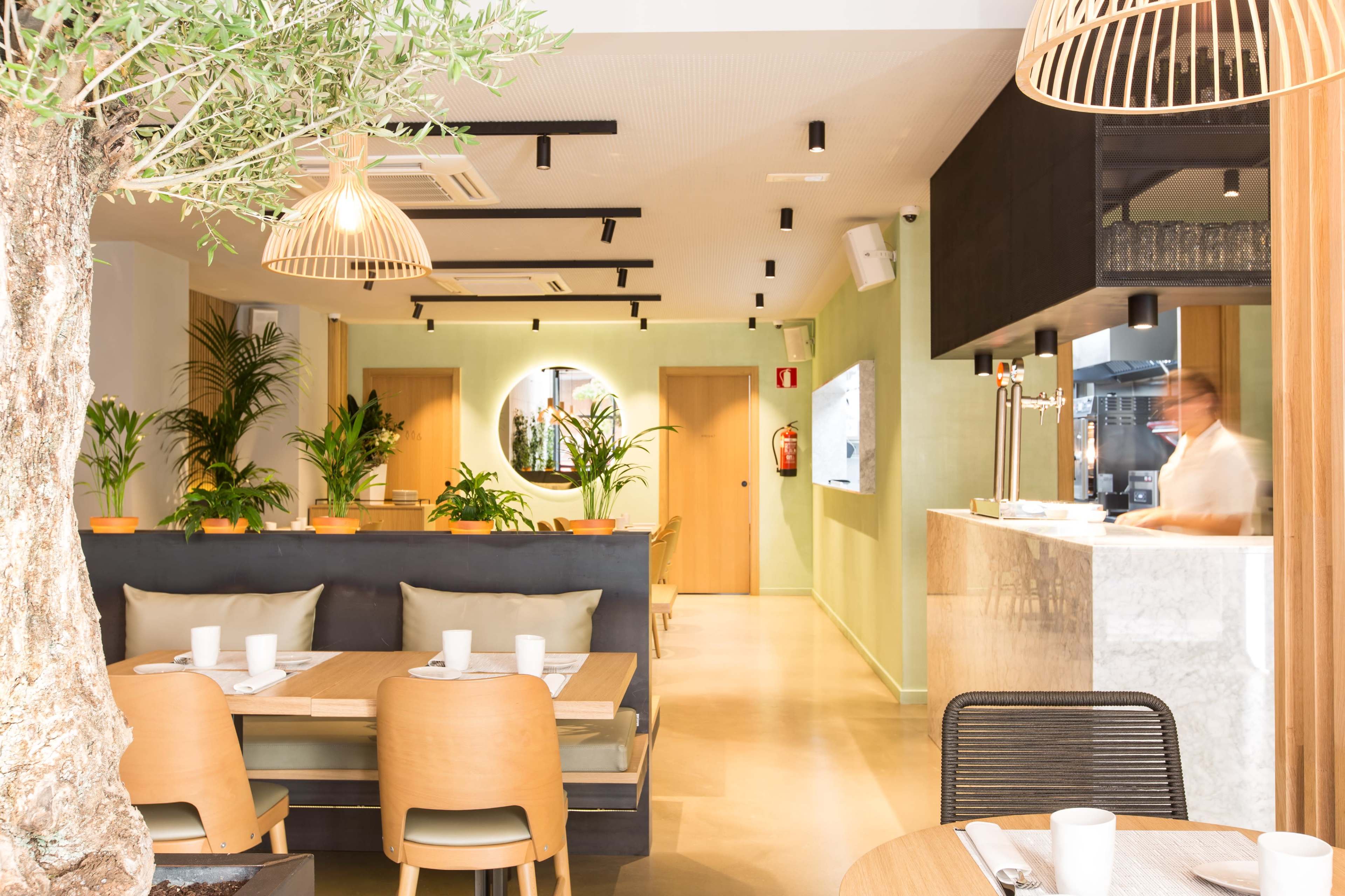 A modern restaurant interior with wooden tables, green plants, and a bar area featuring a marble counter.
