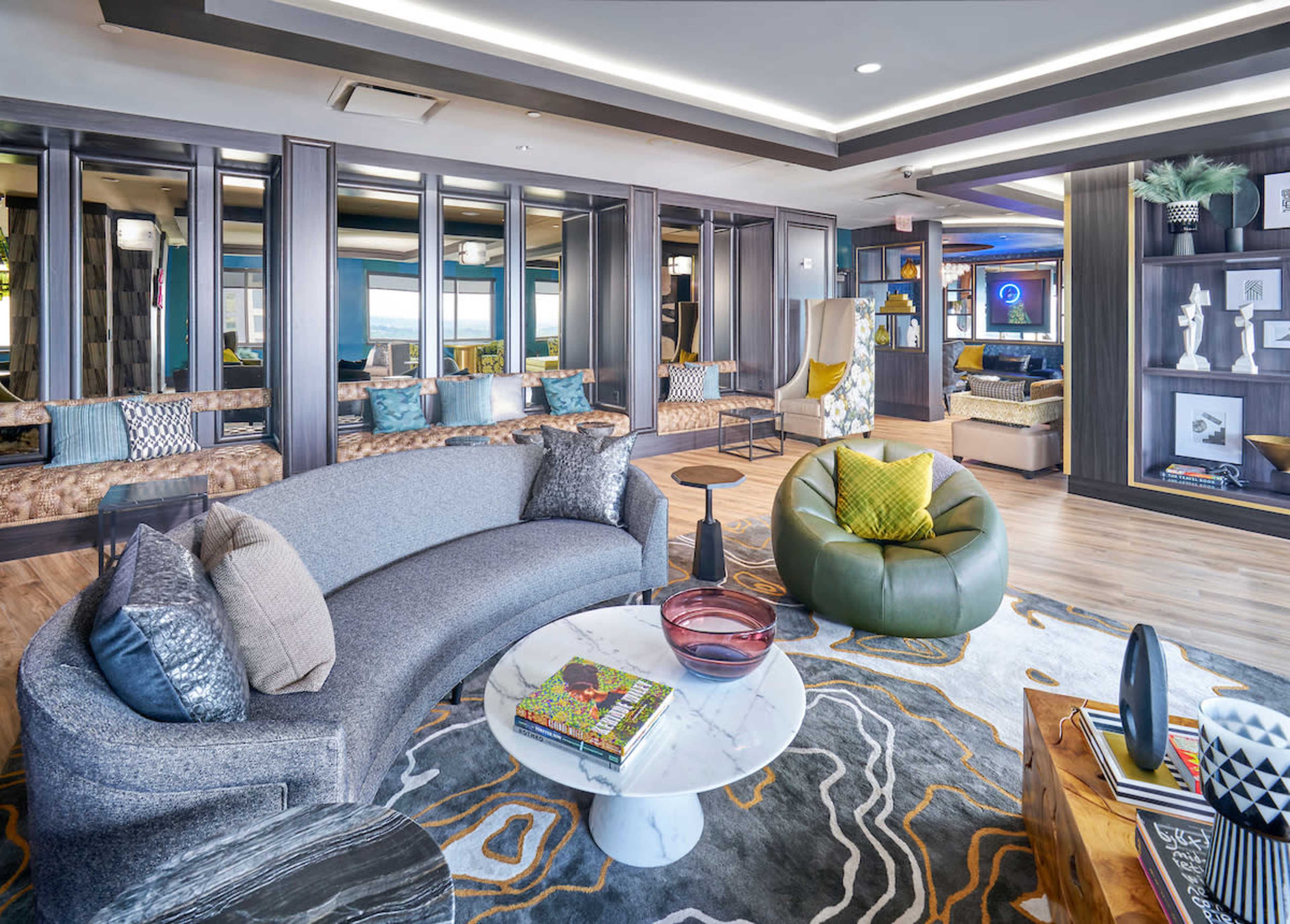 A modern lounge area with a curved gray sofa, a round white coffee table, a green chair, and large windows allowing natural light.
