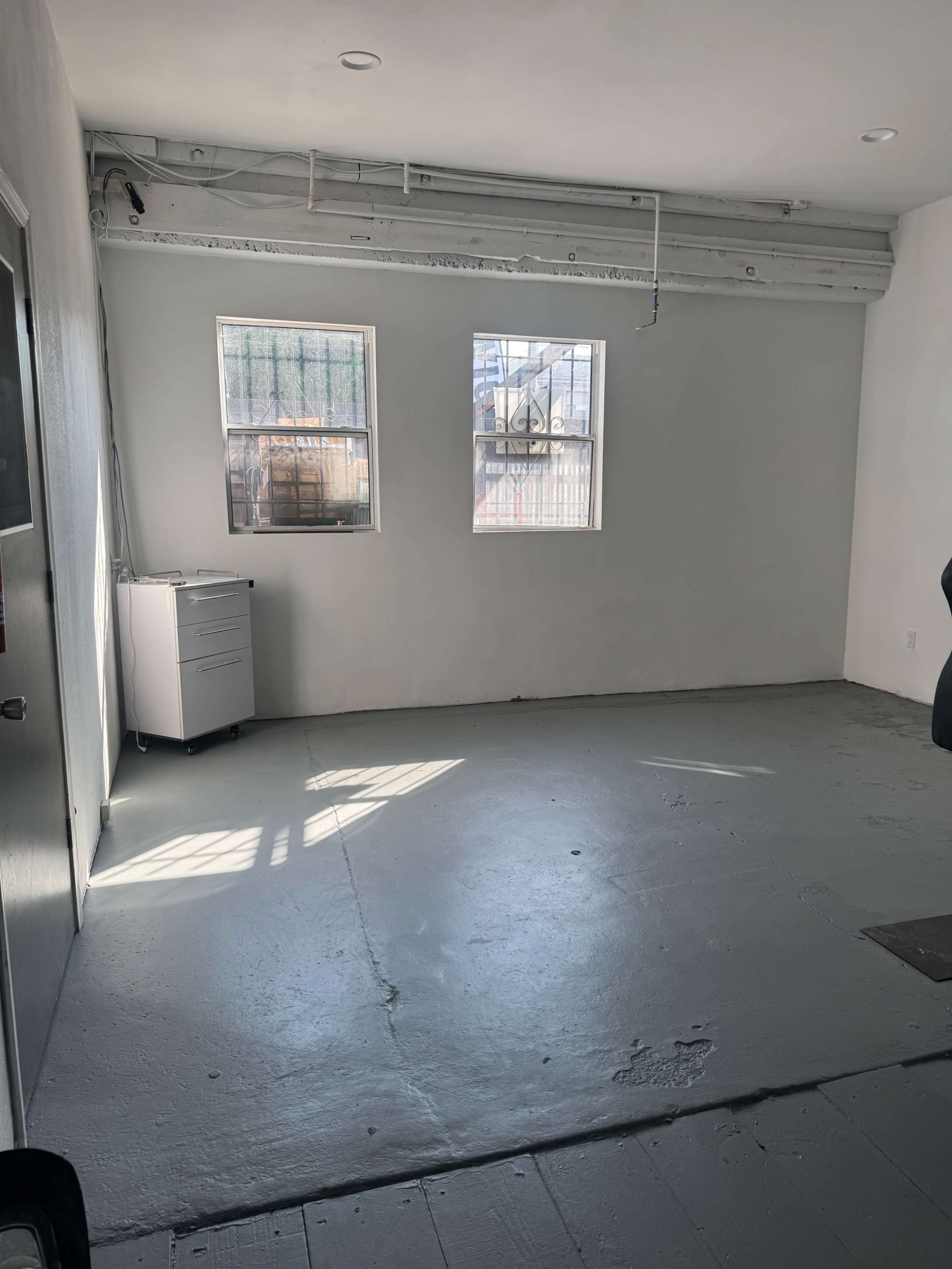 The image shows a sparse, empty room with two windows, a metal filing cabinet, and a bare floor.