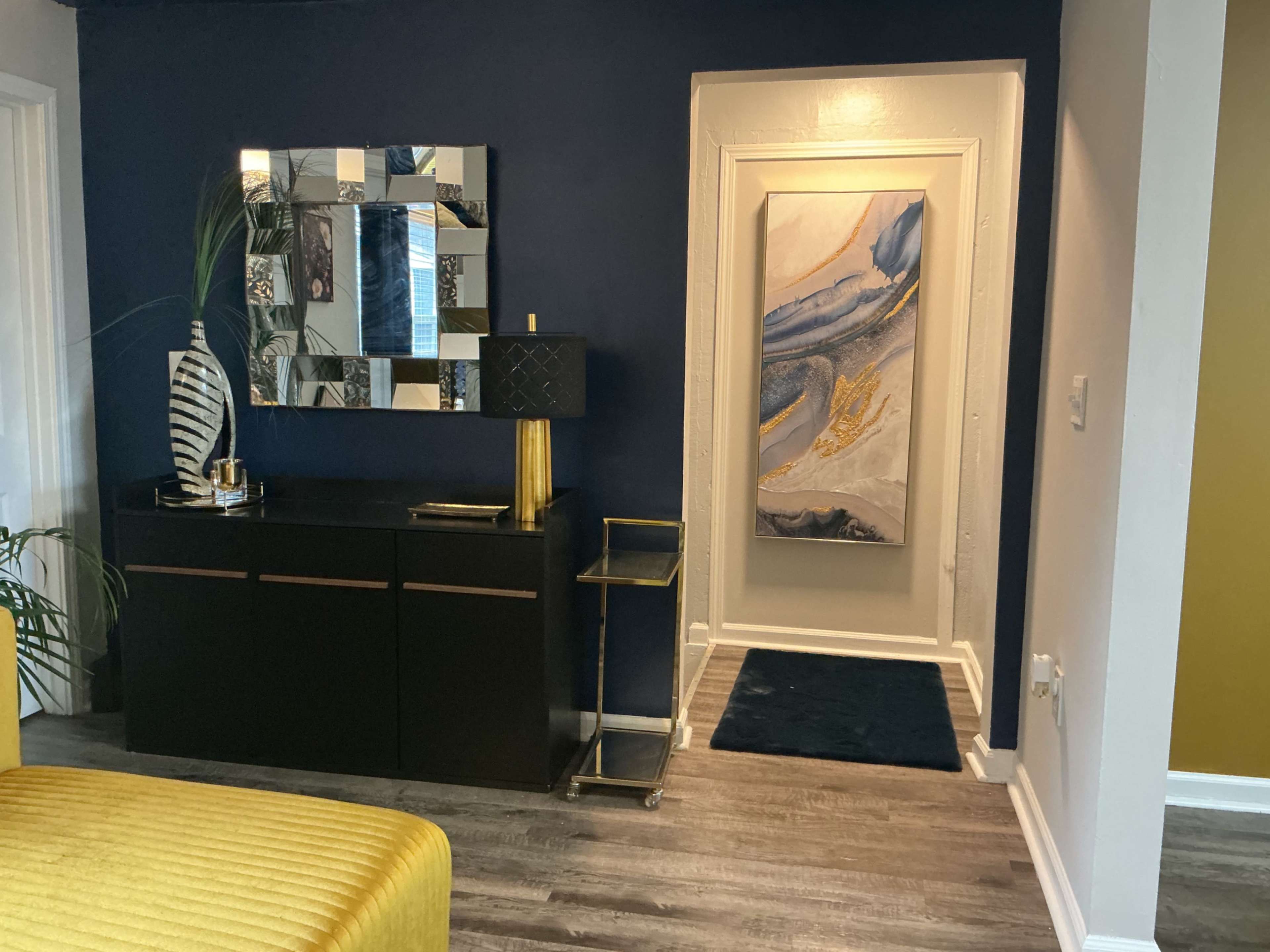 The image shows a hallway with a dark blue wall, a decorative mirror, a black console table with golden accents, and a piece of abstract wall art illuminated by soft light.