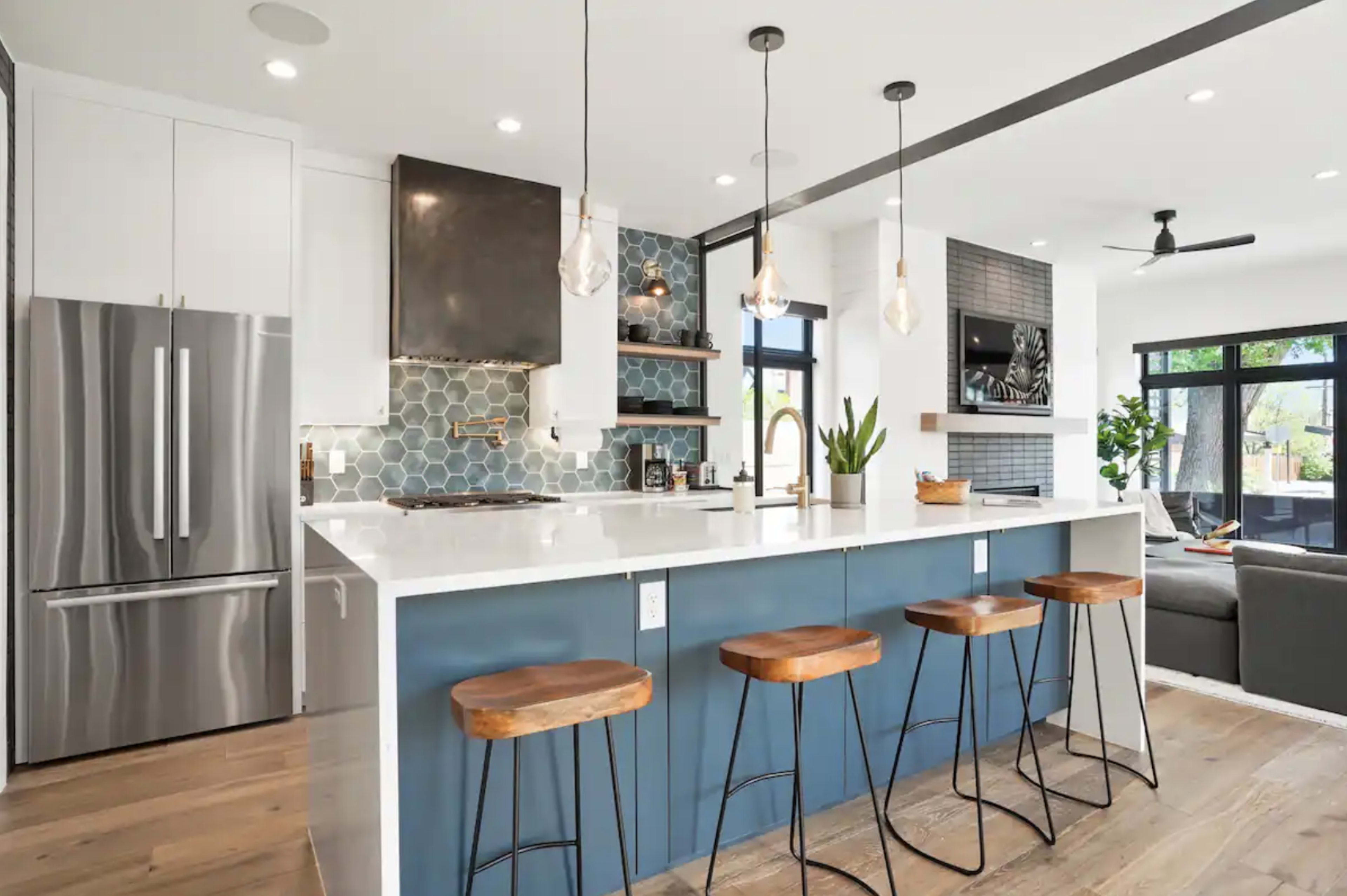 A modern kitchen features a large island with four stools, stainless steel appliances, and a geometric tile backsplash.