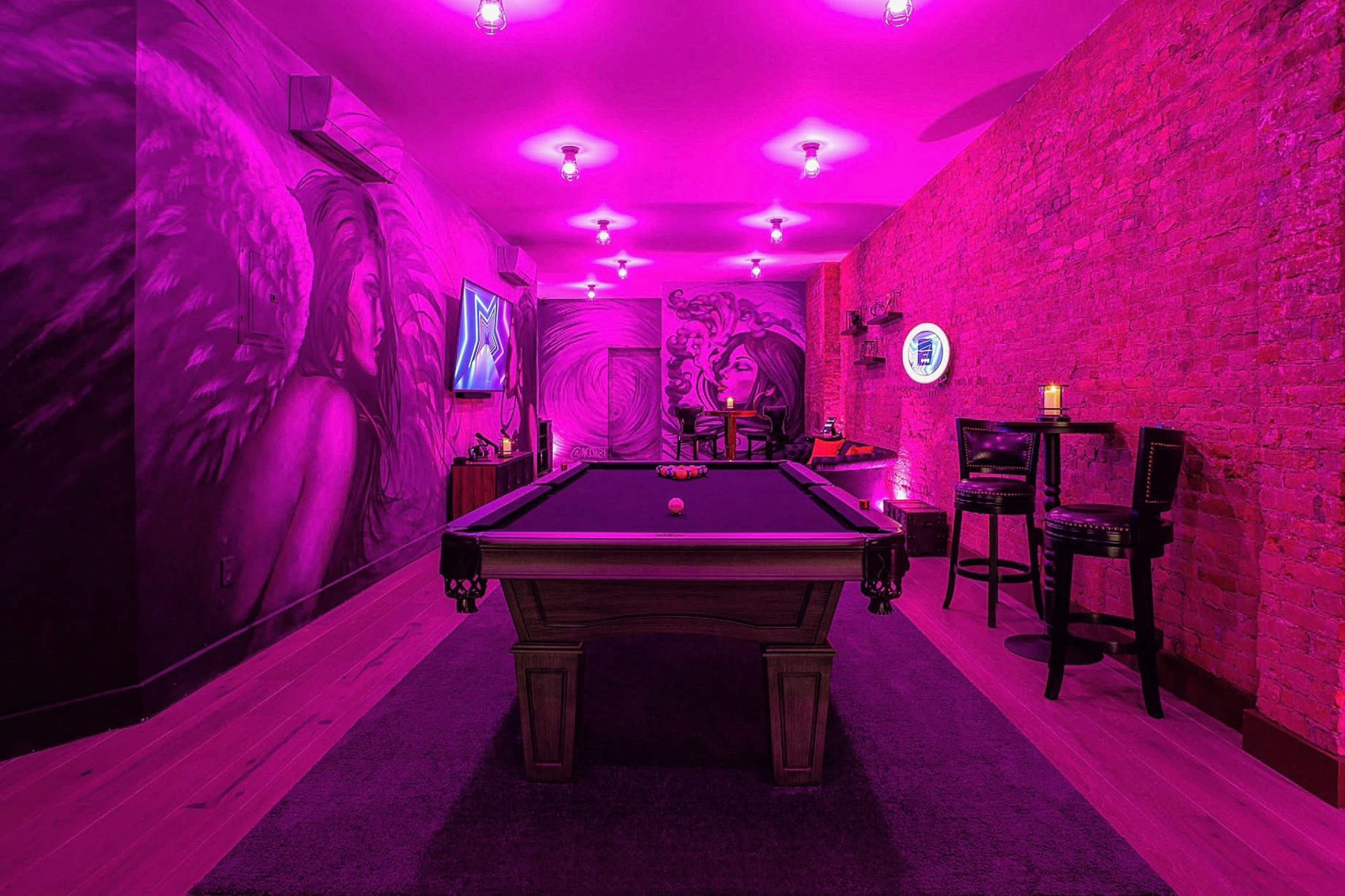 A game room features a billiards table surrounded by brick walls painted with vibrant murals and illuminated by pink lights.