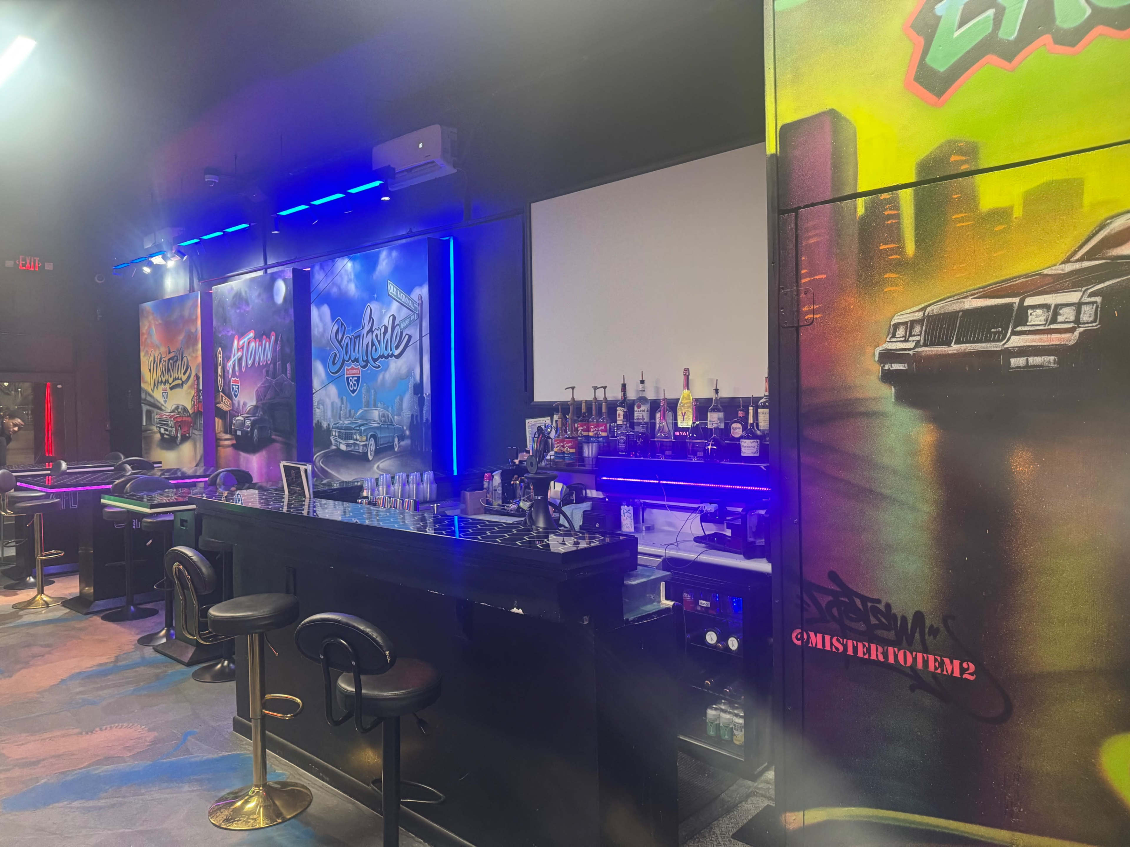 The image shows a bar setup featuring vibrant graffiti murals on the walls and a backlit countertop with various bottles lined up.