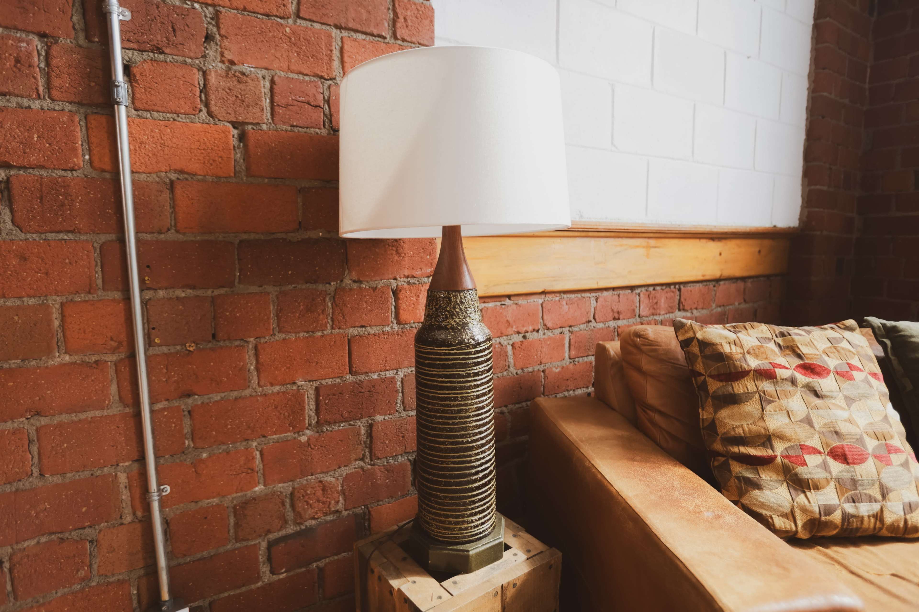 A vintage lamp with a textured base and a white shade stands beside a brown leather sofa against a brick wall.