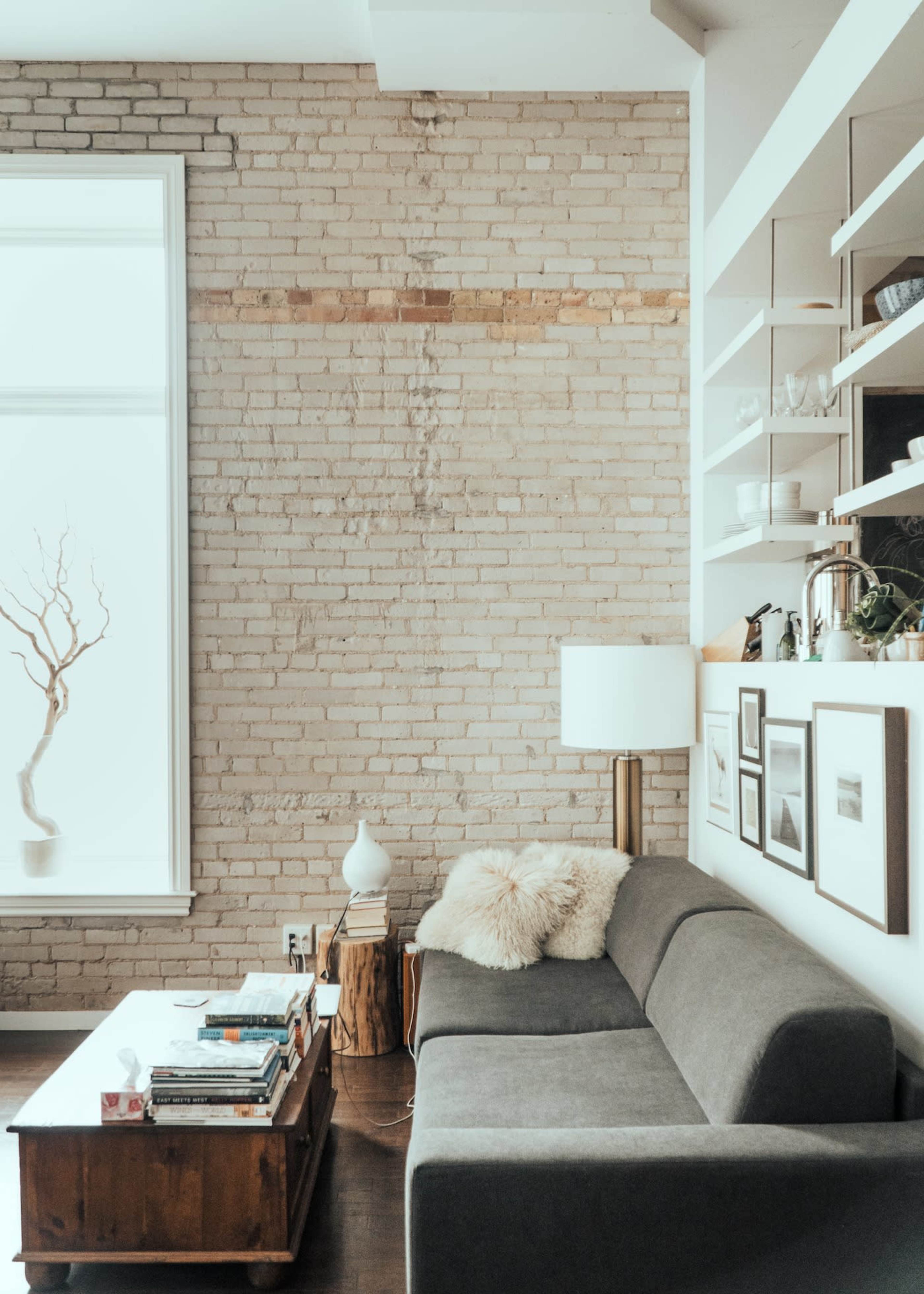 A cozy living room with a gray sofa, a wooden coffee table stacked with books, and a large window with natural light illuminating a brick wall.