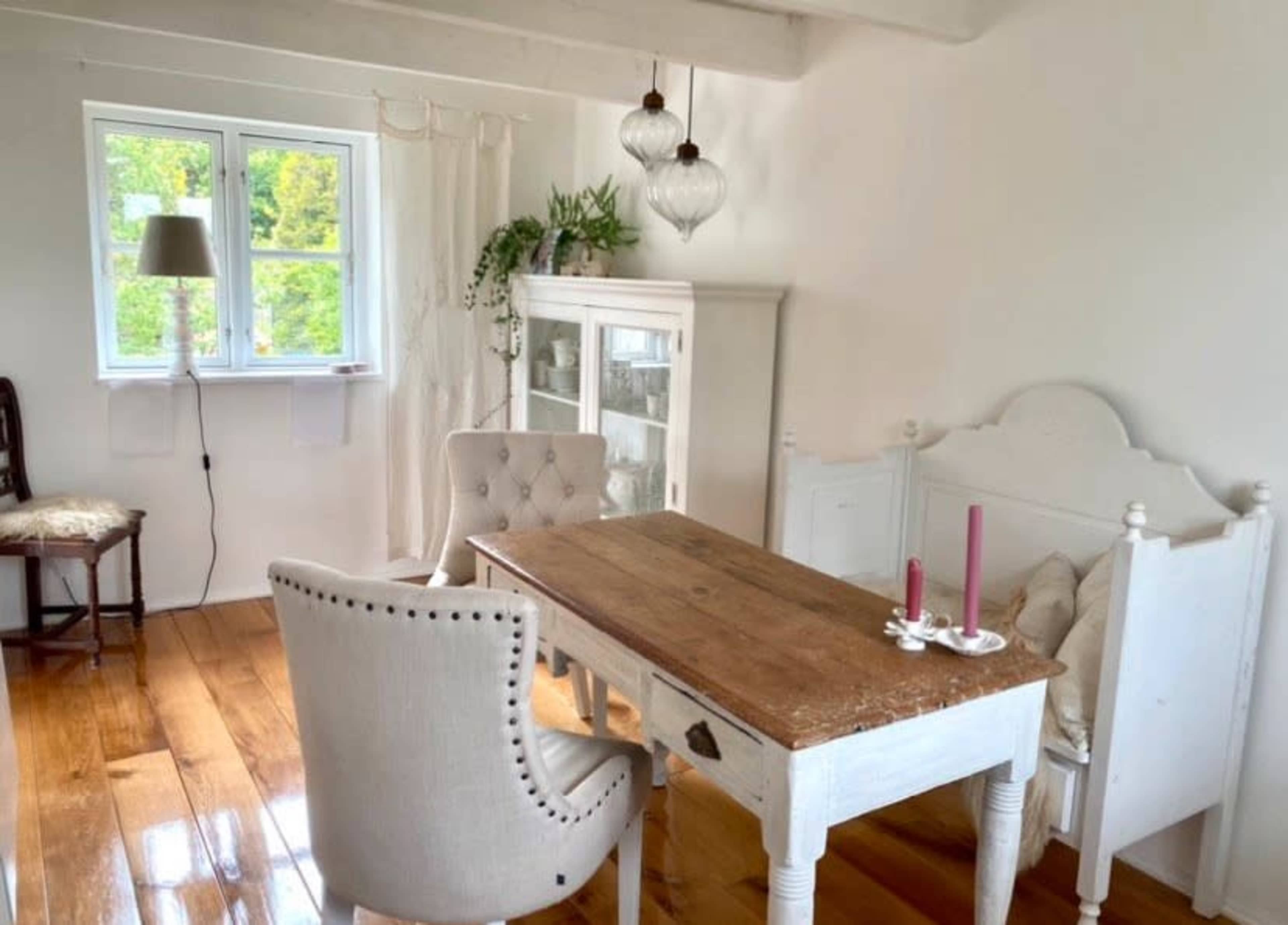 The image shows a bright, minimalistic dining area featuring a wooden table surrounded by two upholstered chairs, a glass cabinet, and hanging light fixtures.