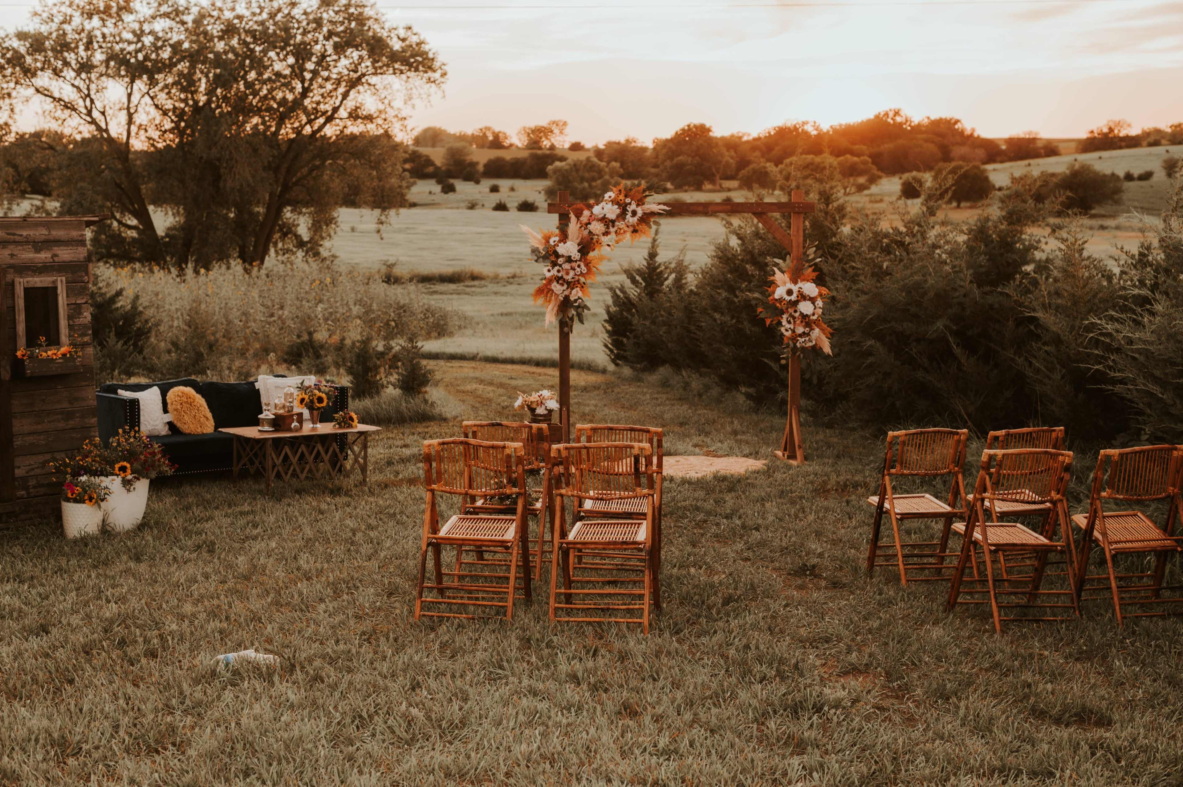 The scene features an outdoor wedding setup with wooden chairs arranged around a decorative arch adorned with flowers, overlooking a grassy field at sunset.