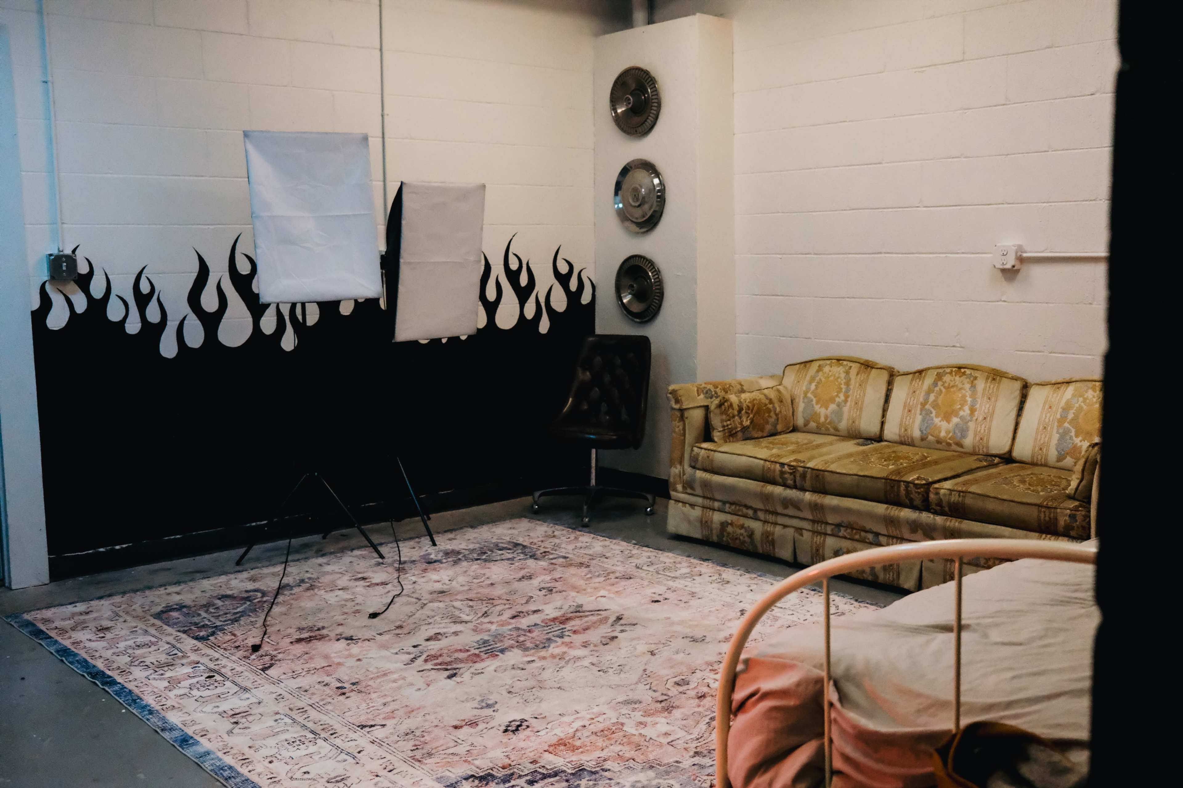 The image shows a small room with two white backdrops on stands, a patterned rug, a vintage-style couch, and a black chair, all set against a wall with flame designs.