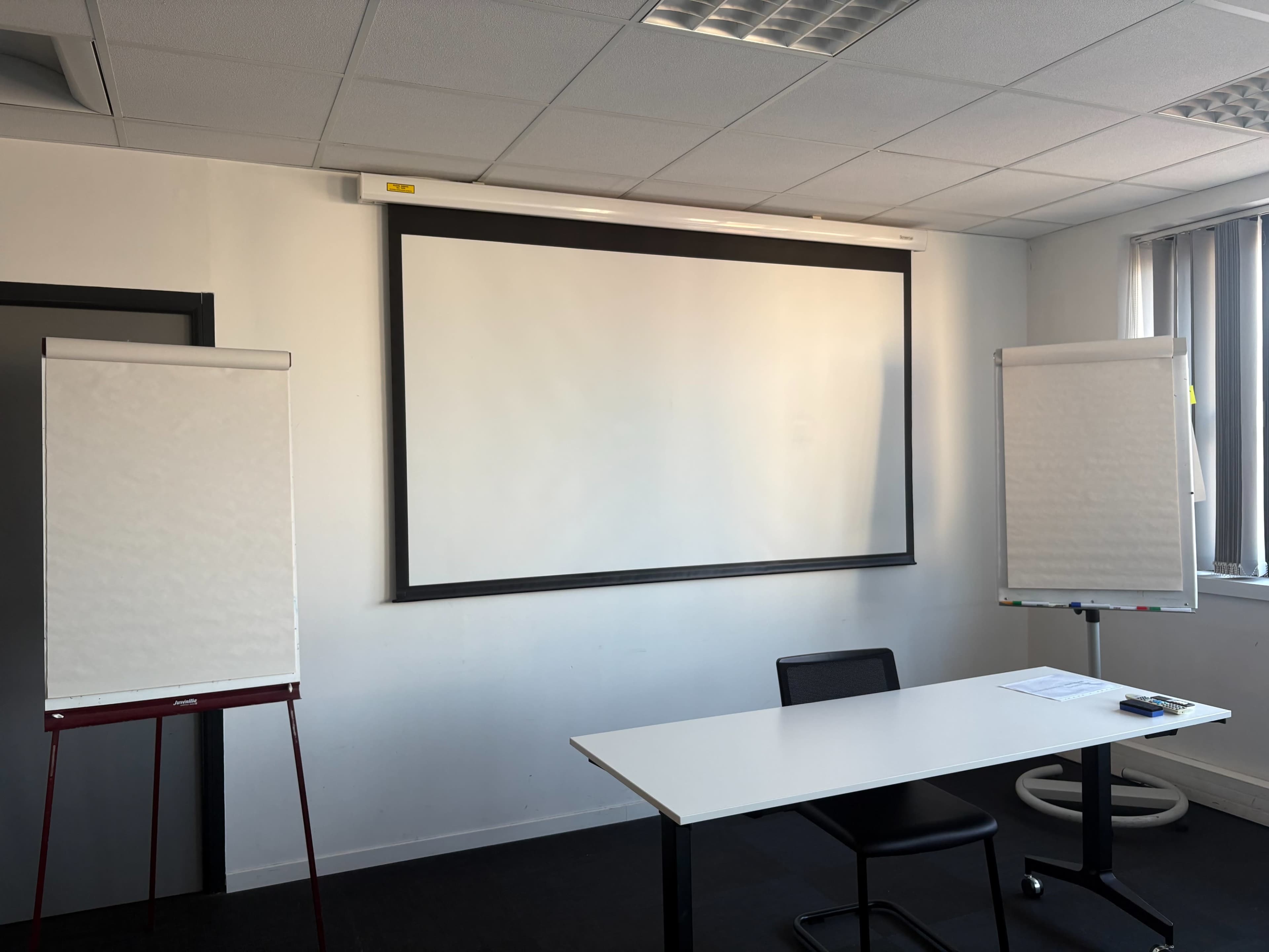 The image shows a meeting room with a blank projector screen, a white table and chair, and two flip charts on stands.