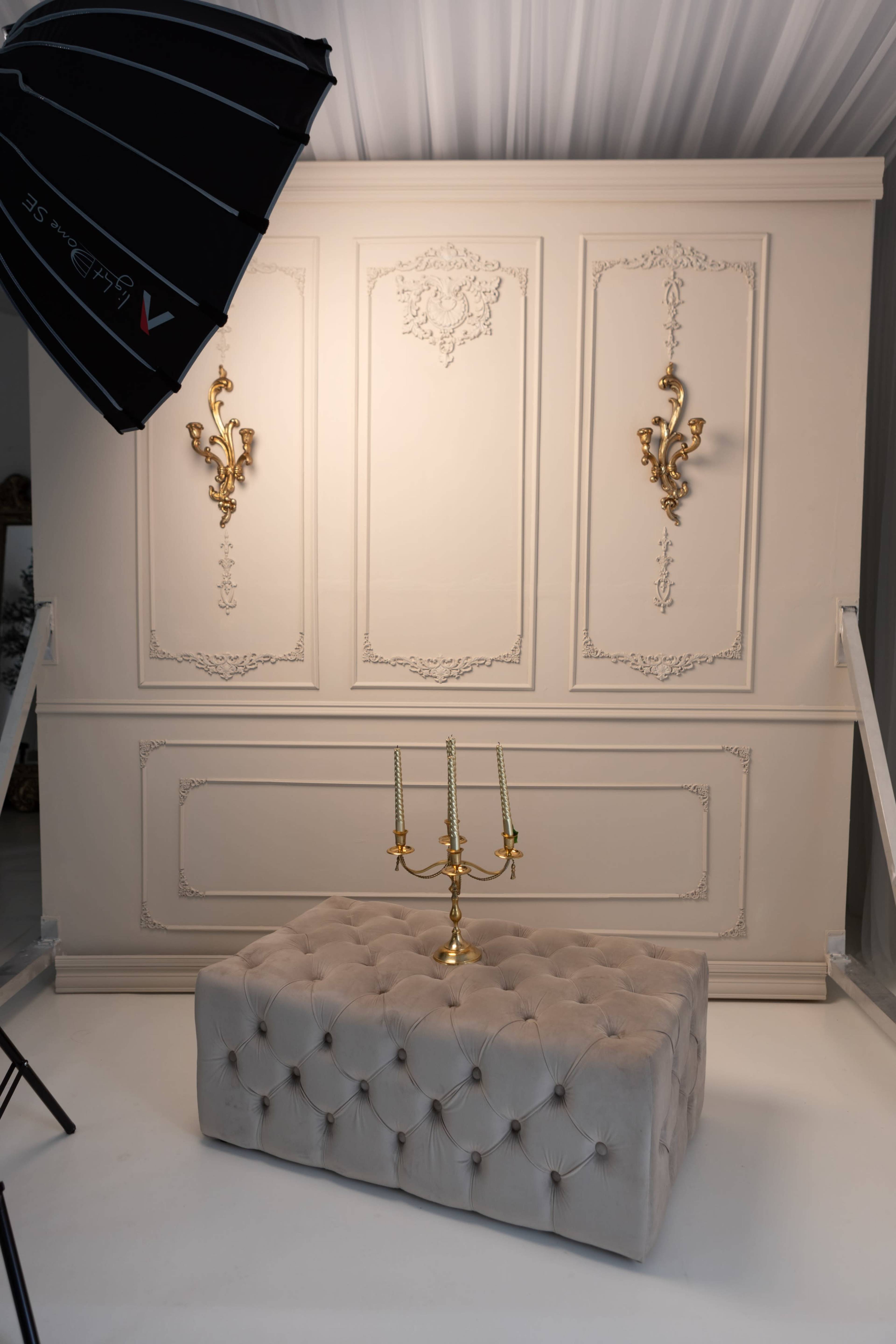 A tufted beige ottoman sits in front of an ornate white wall adorned with decorative sconces and classical designs, illuminated by a large photography umbrella.