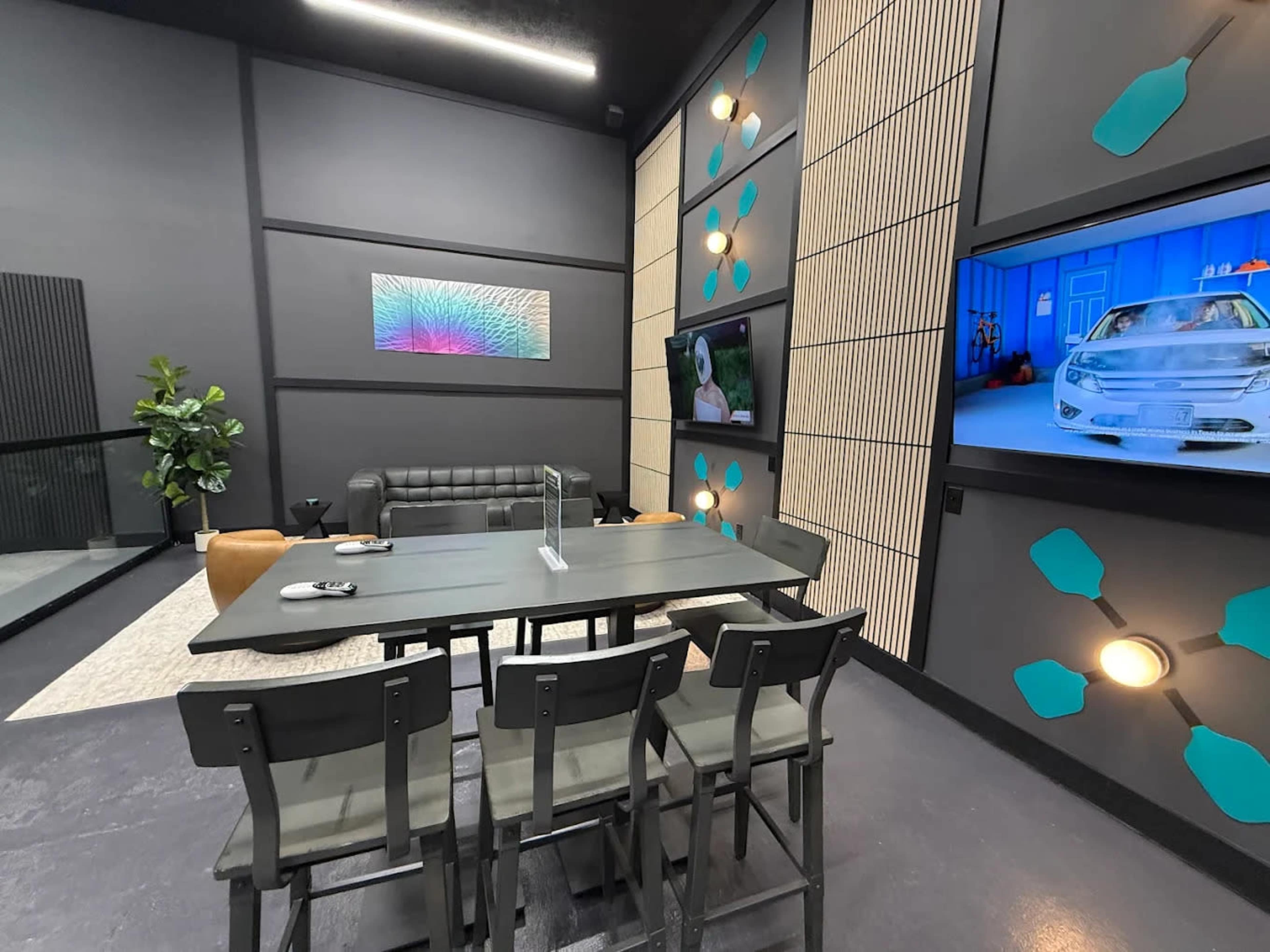 The image shows a modern meeting room with a dark wall design, a large table surrounded by six chairs, a black leather couch, and two screens displaying images.