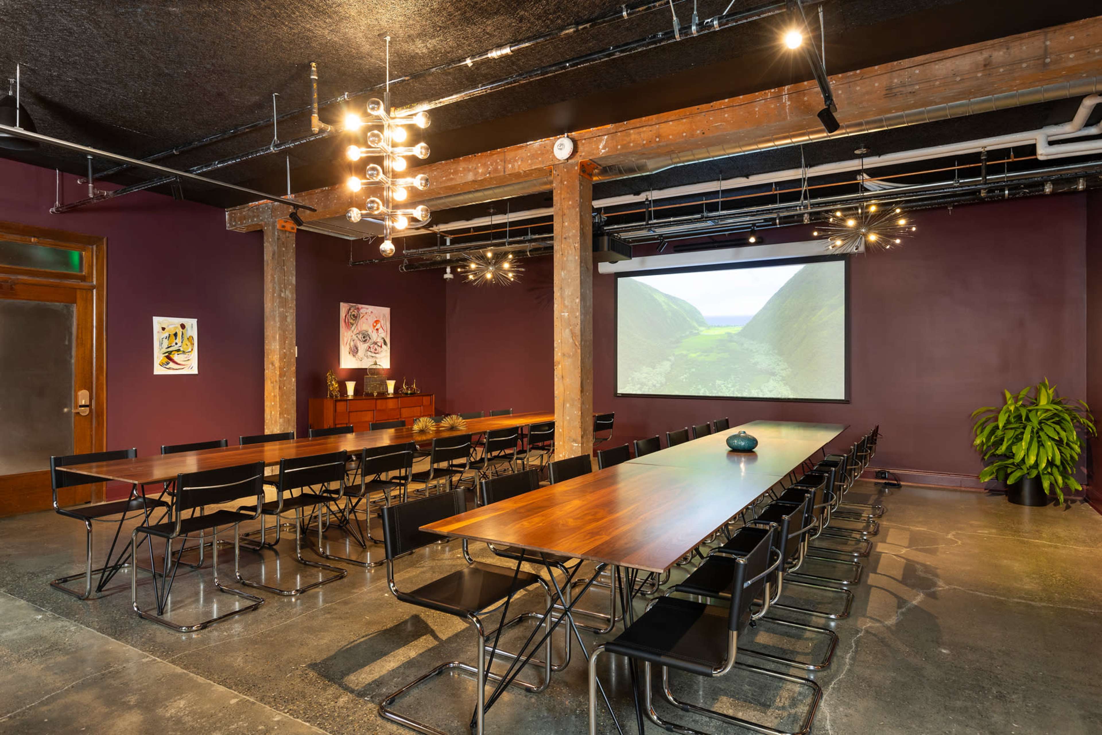 A modern conference room with a long wooden table, black chairs arranged in rows, and a large projector screen displaying a scenic landscape.