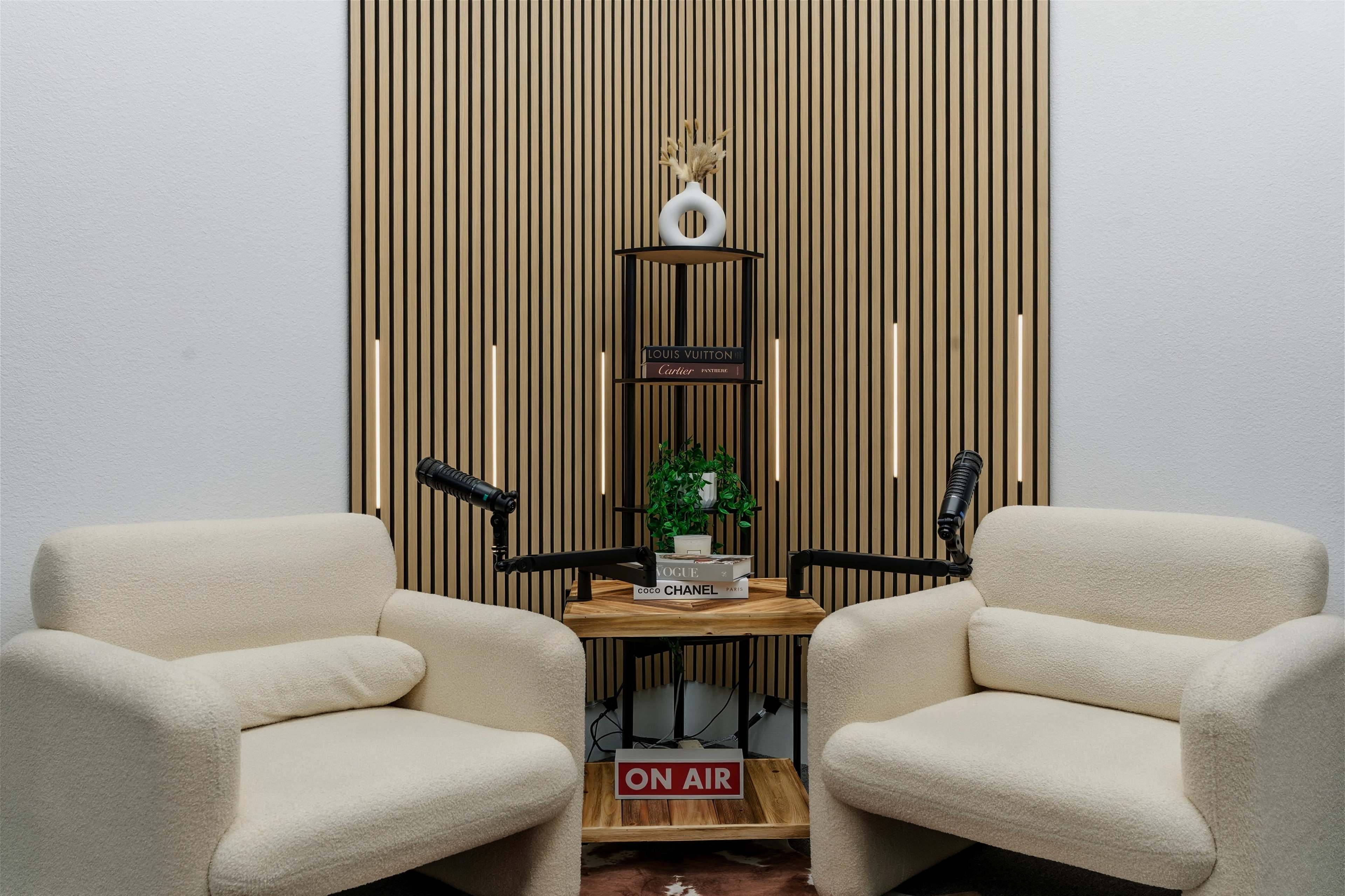 The image features two cream-colored armchairs facing a wooden shelf with a small plant and decorative items, set against a backdrop of vertical wooden slats.