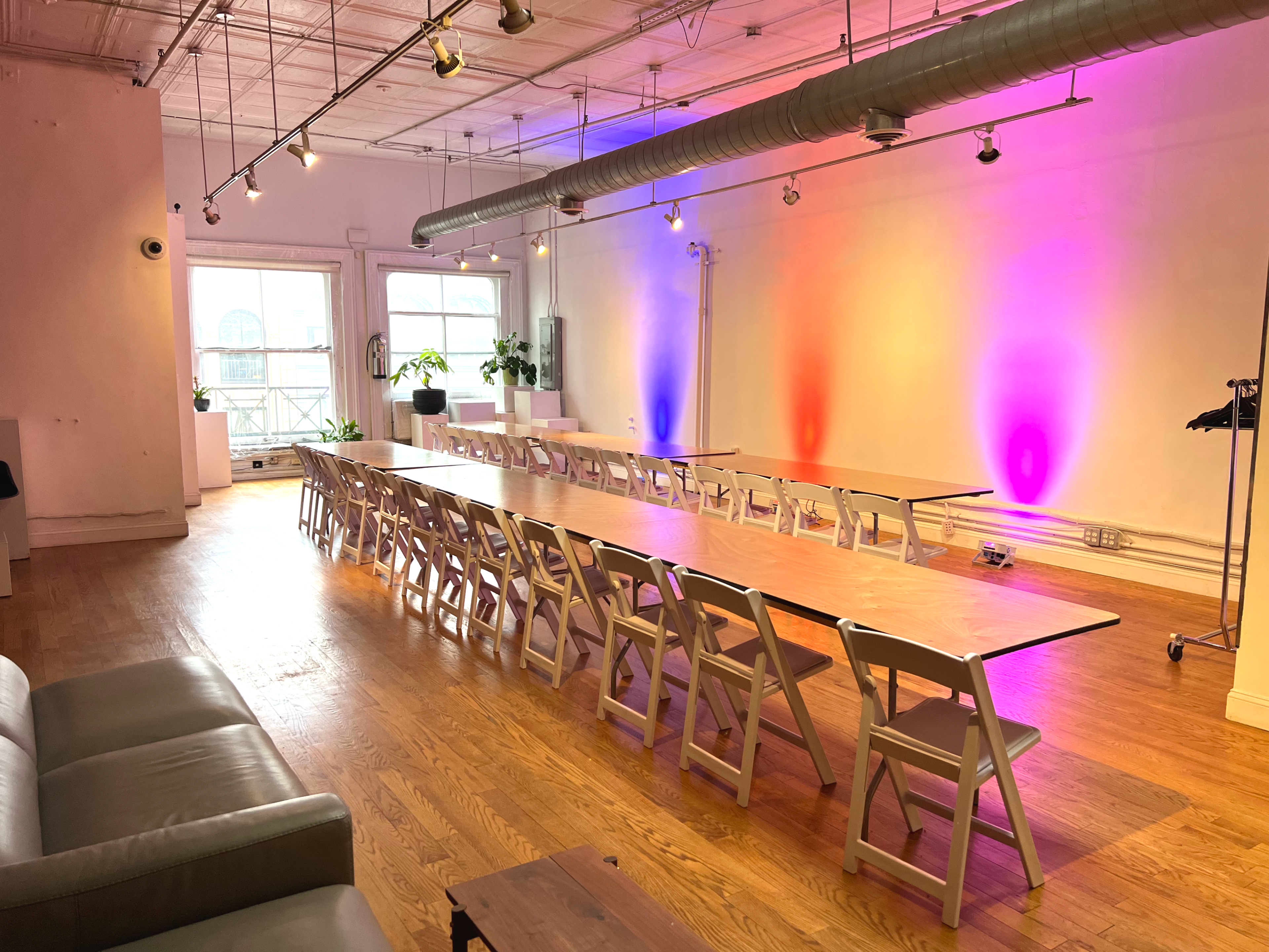 A long wooden table with folding chairs is set up in a bright, spacious room featuring colorful lighting and large windows.
