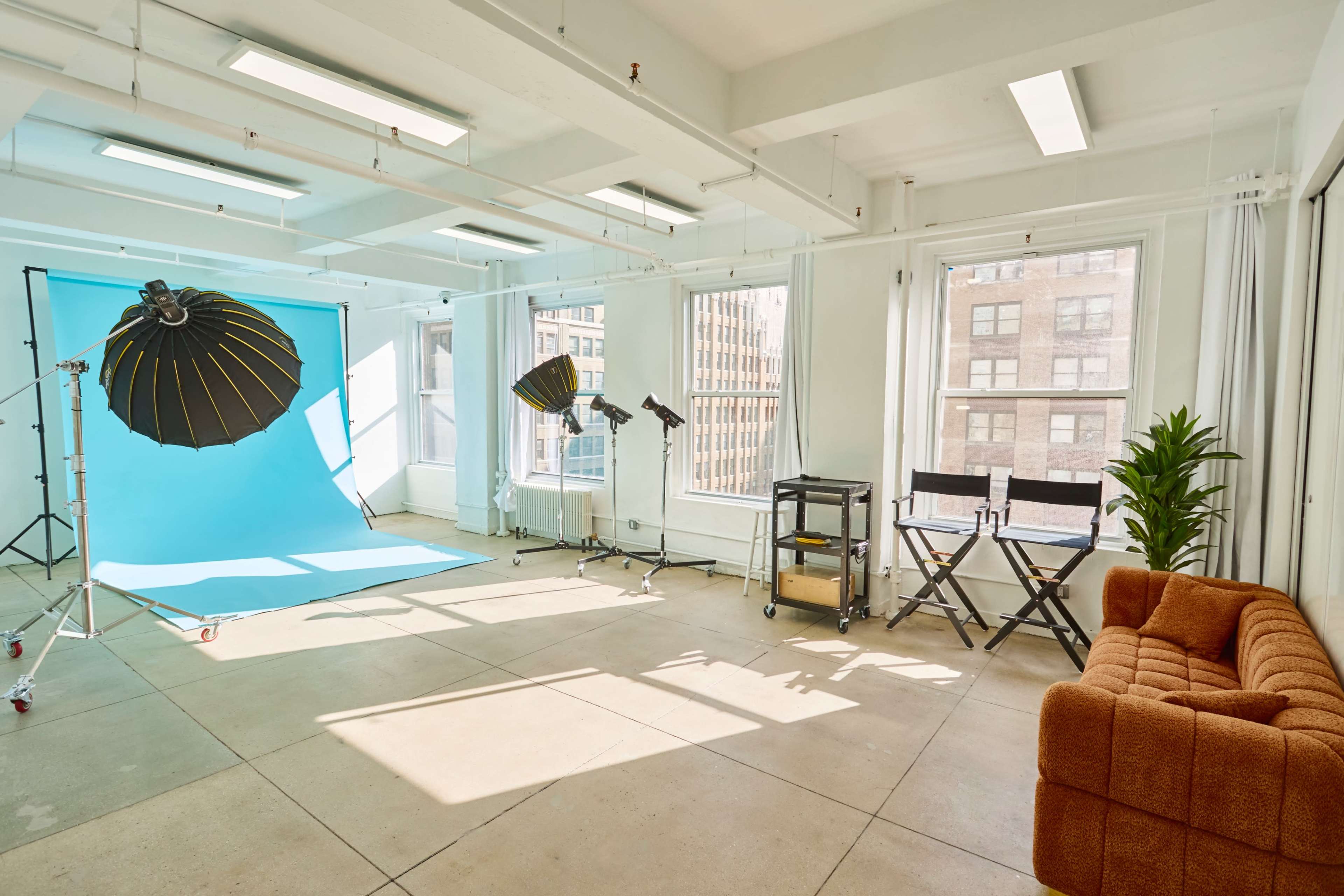 The image shows a photography studio with a bright blue backdrop, lighting equipment, two director's chairs, and a brown couch, all arranged in a spacious, well-lit room with large windows.