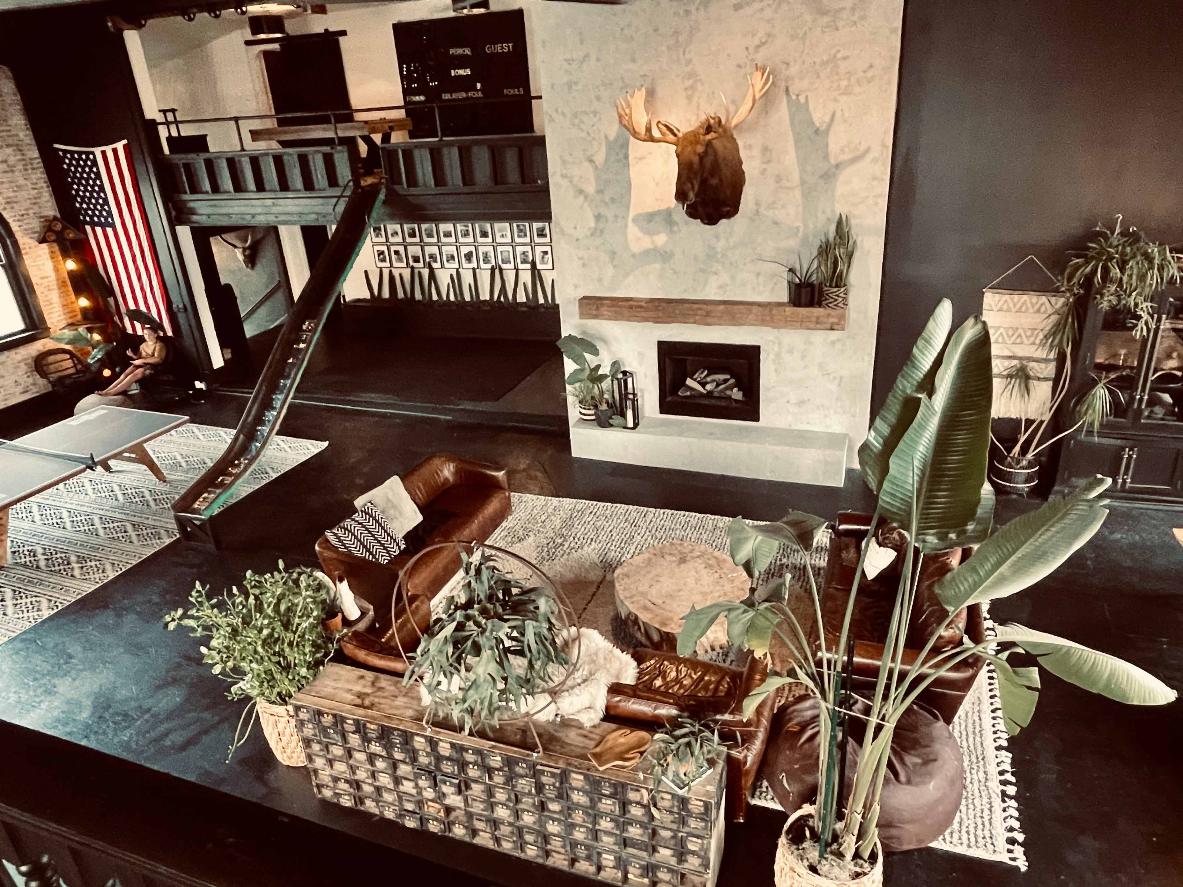A spacious room with a mix of modern and rustic decor, featuring a large seating area with plants, a wall-mounted moose head, and an American flag in the background.