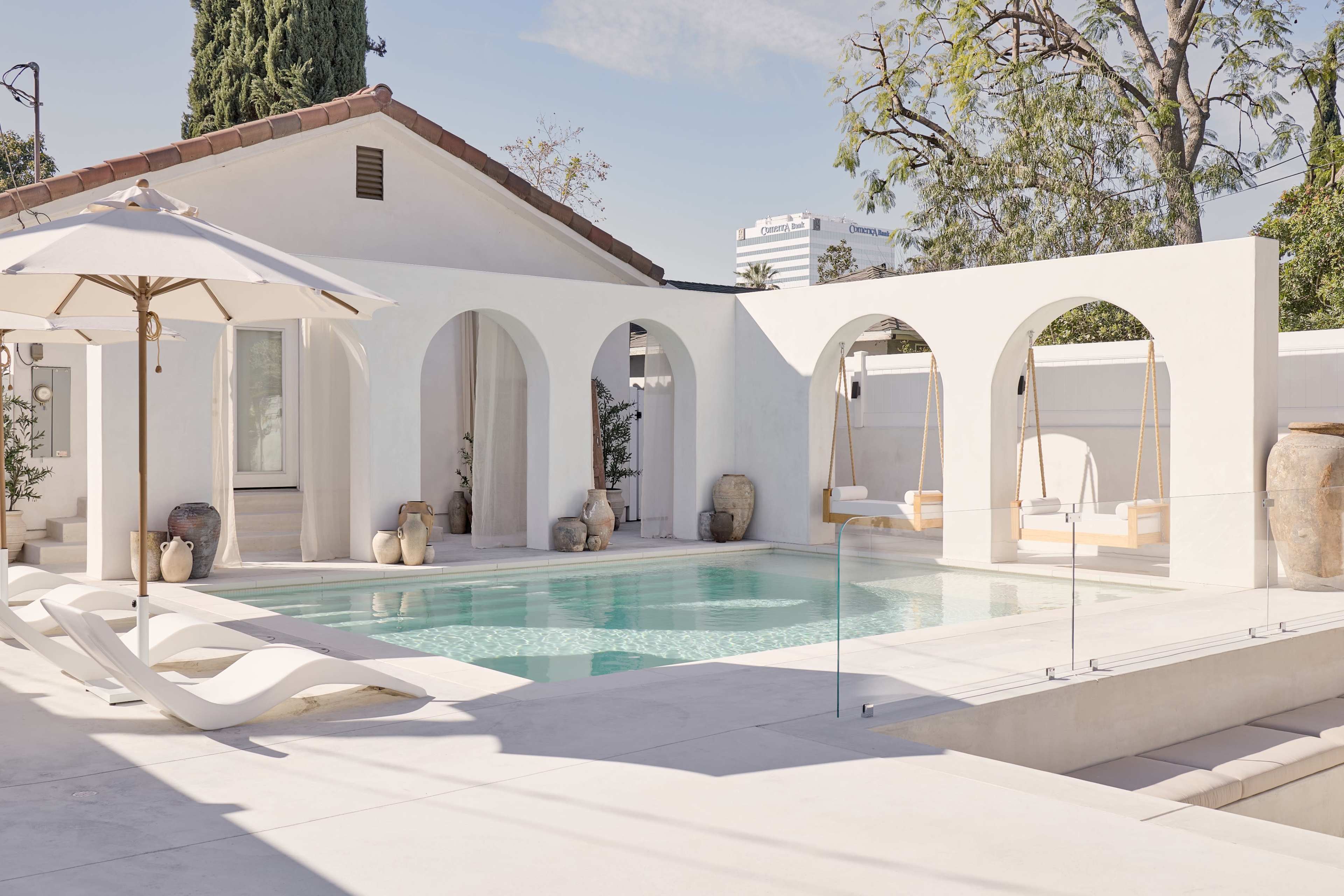 The image shows a modern outdoor space with a swimming pool, white walls, archways, and relaxing swing chairs.