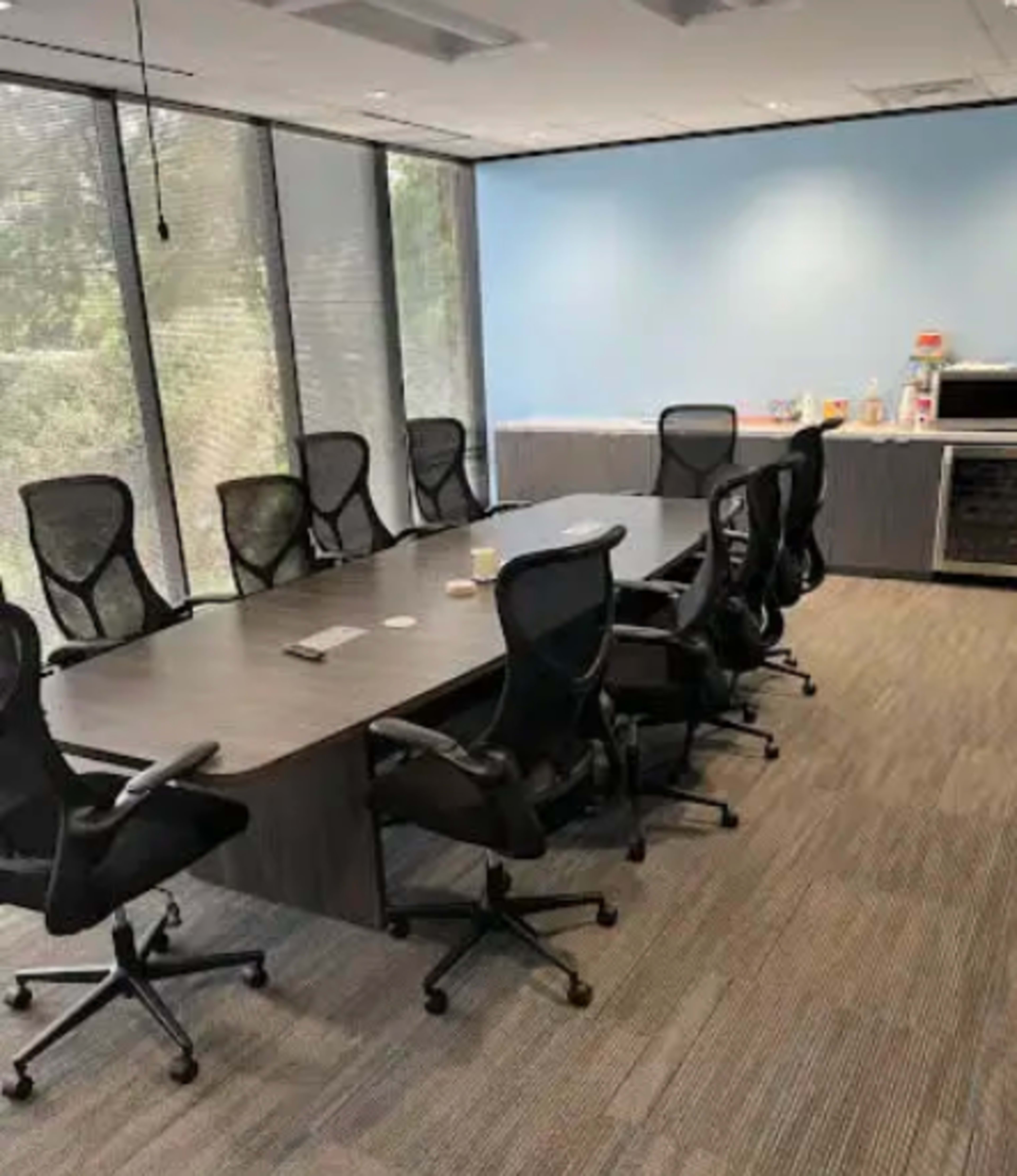 The image shows a conference room with a long table surrounded by eight black mesh office chairs, along with a counter area featuring some supplies and a window view.