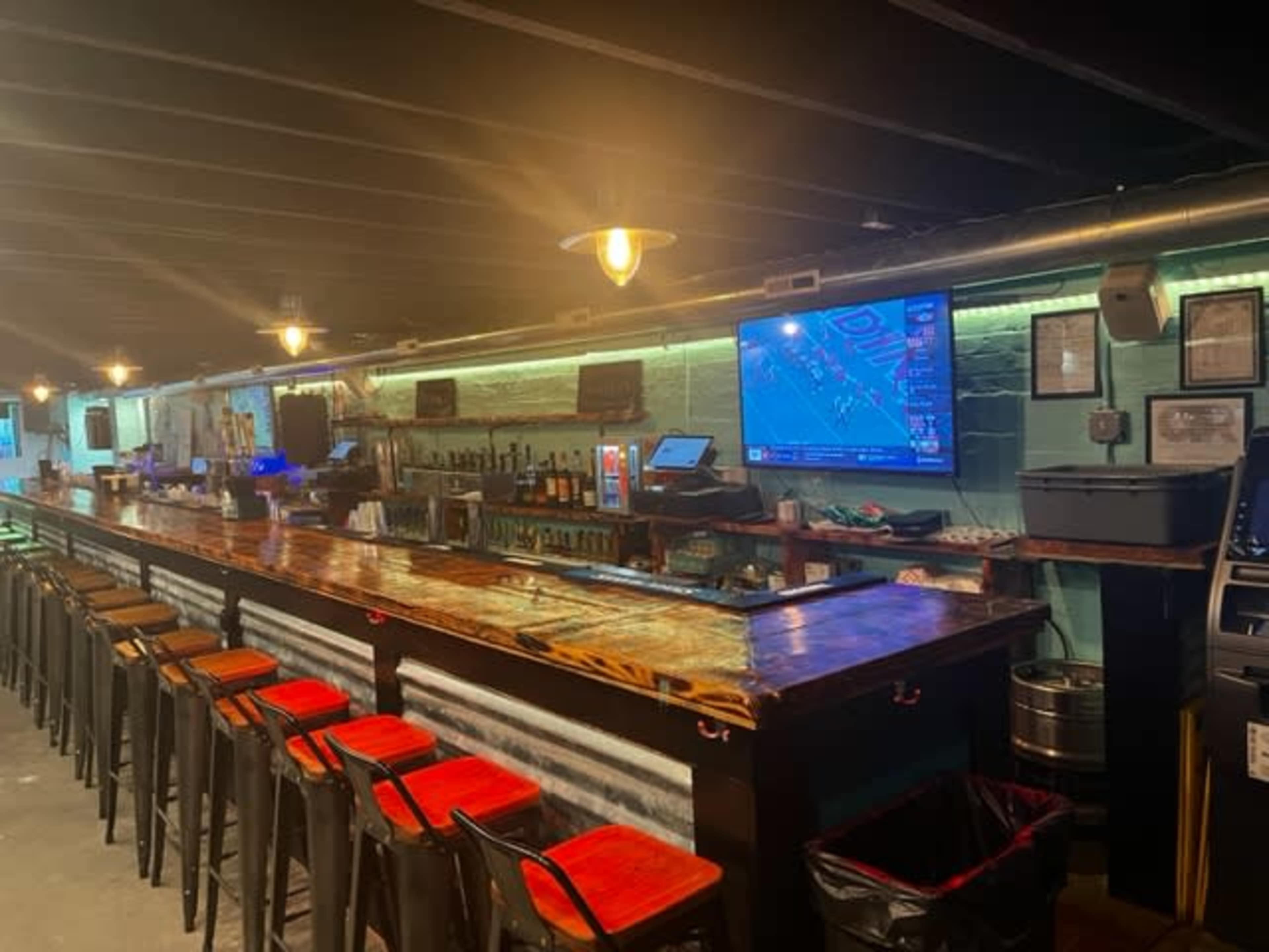 A dimly lit bar with a long wooden countertop, several bar stools, and a television displaying a sports game on the wall.