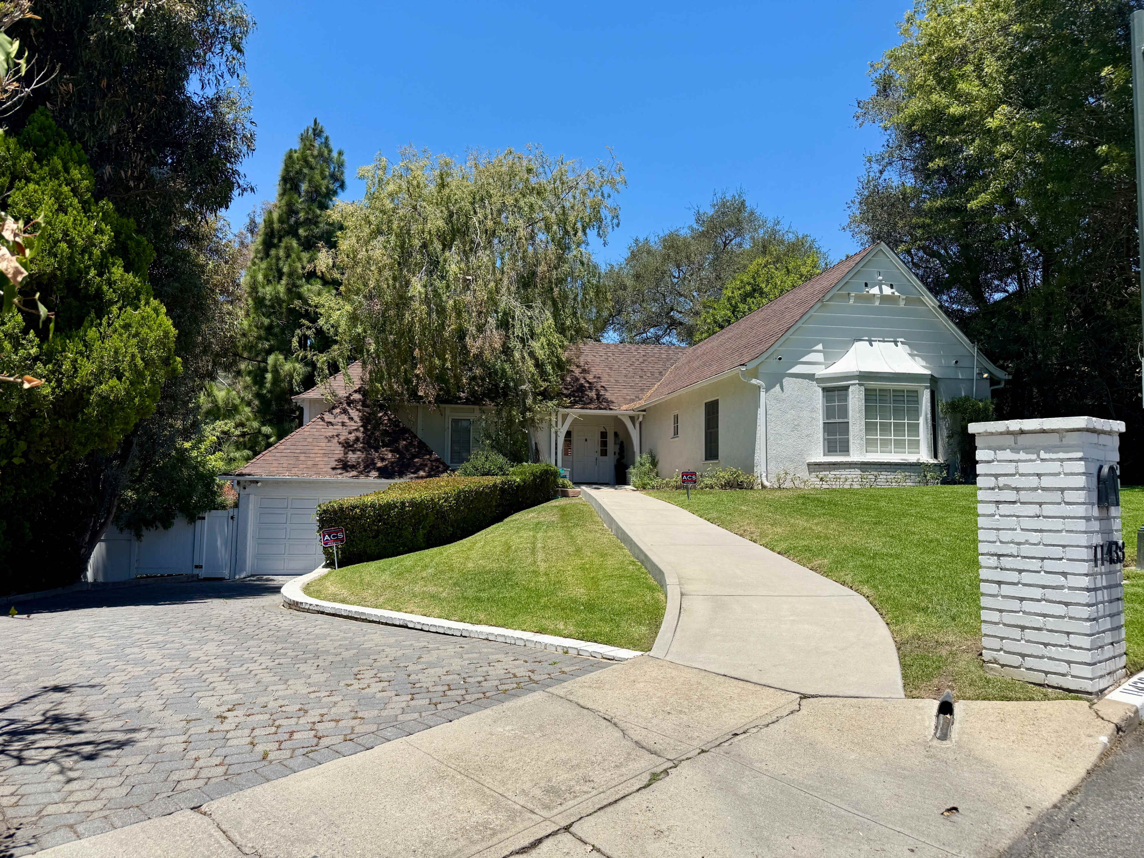 The image shows a single-story house with a large front yard, a curved driveway, and several trees surrounding the property.