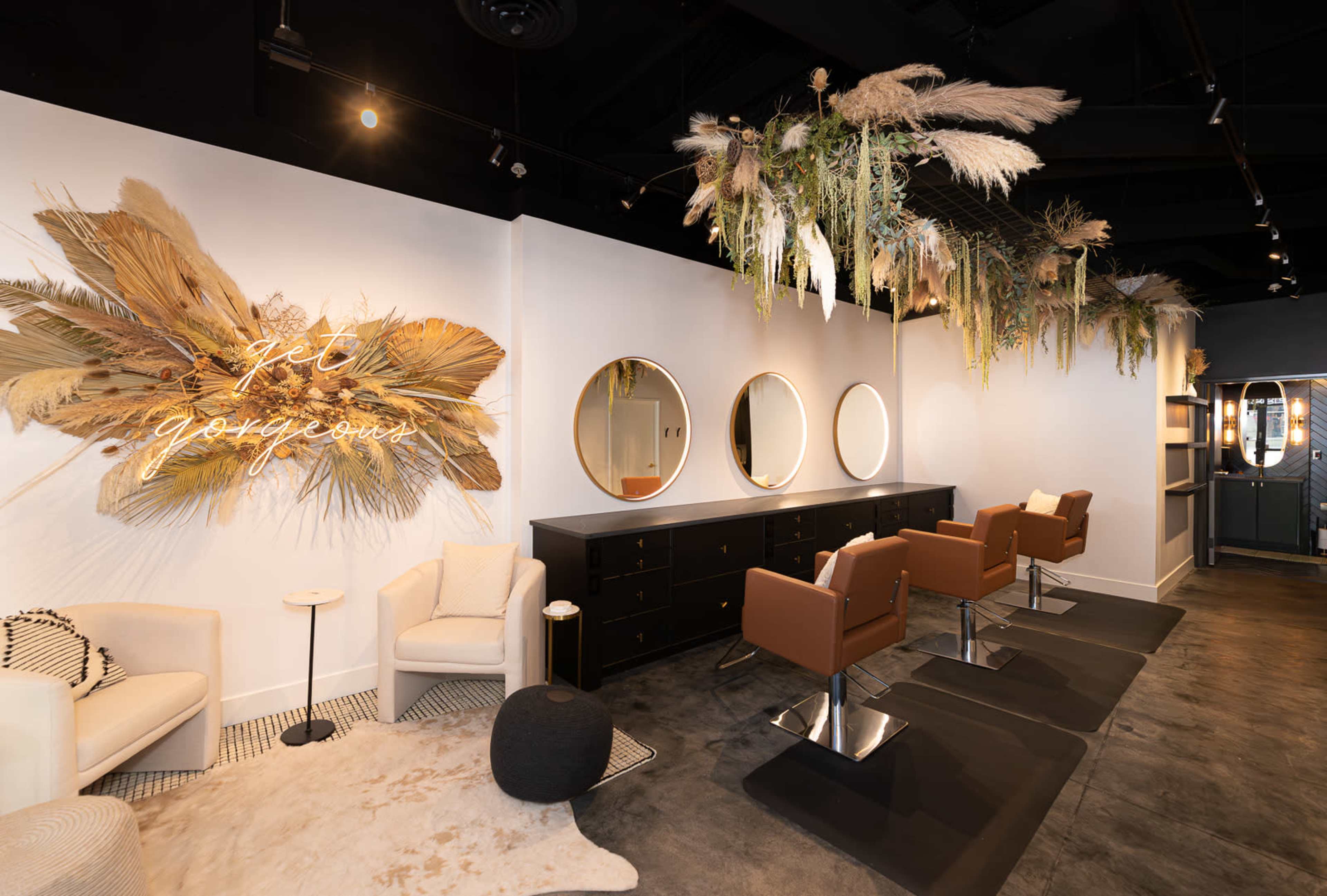 The image shows a modern beauty salon with a stylish interior featuring round mirrors, brown chairs, and greenery decor hanging from the ceiling.