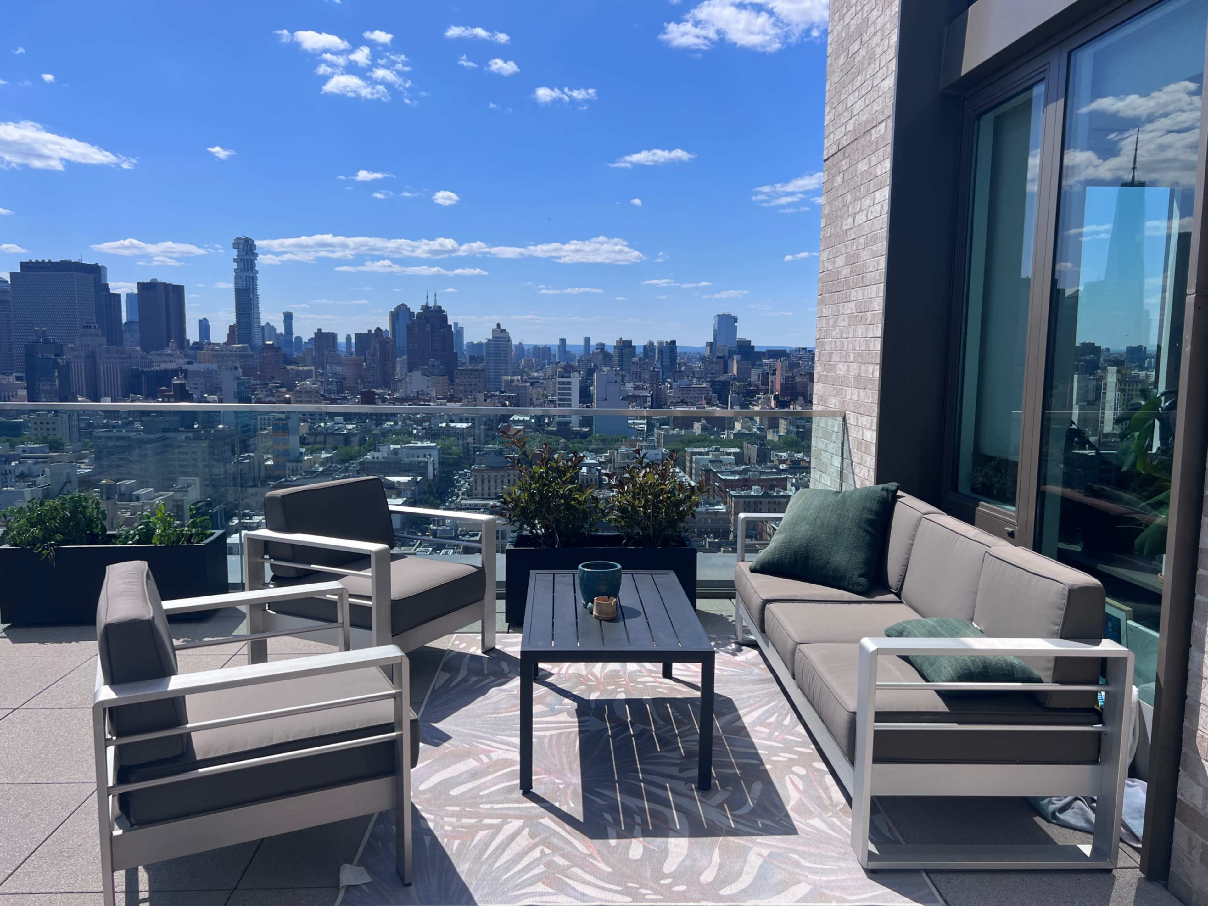 A modern outdoor seating area with gray sofas and a glass table offers a view of the city skyline under a partly cloudy sky.