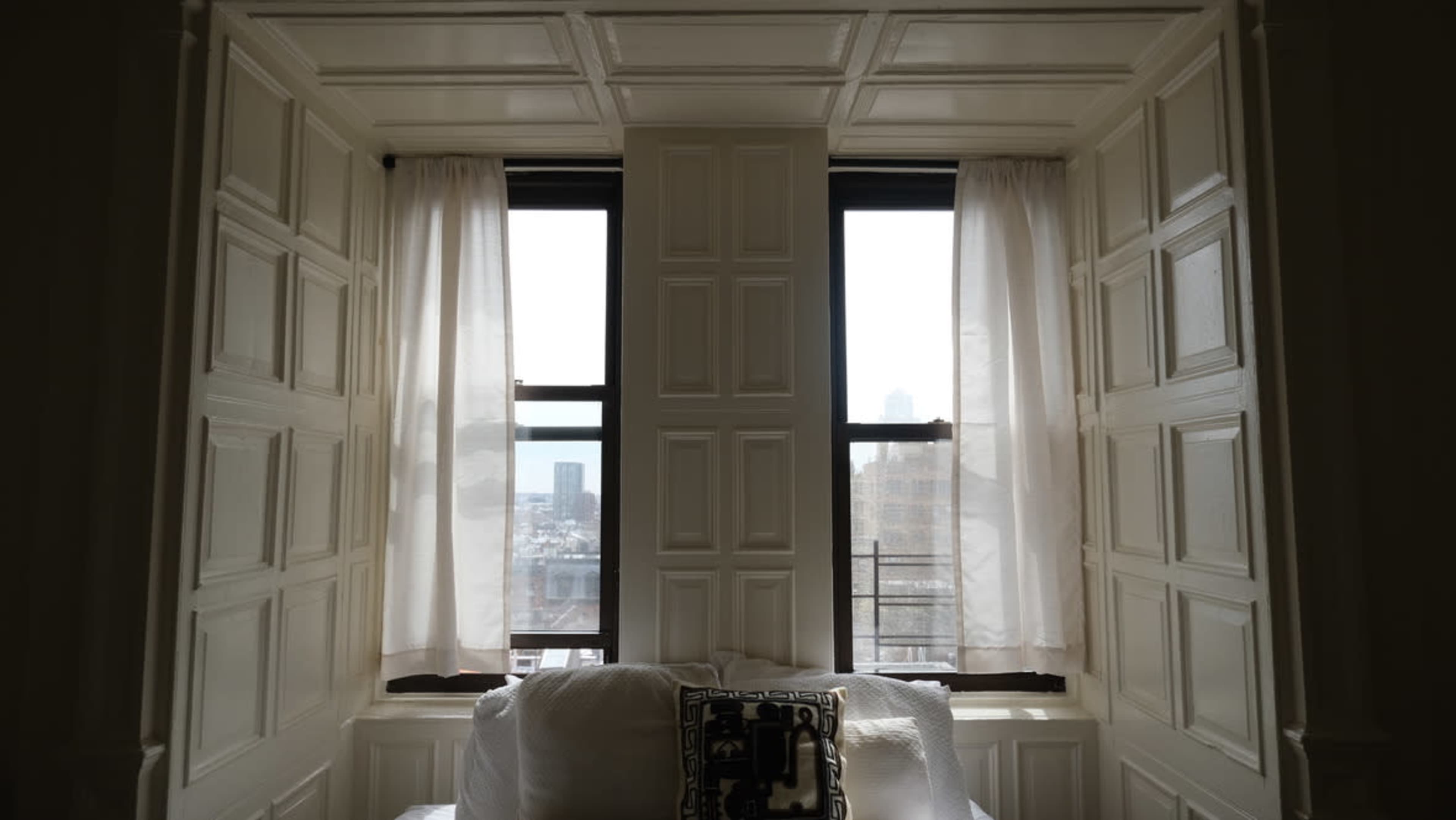 A white-paneled window with sheer curtains and a view of a city skyline in the background, framed by a light-colored wall.