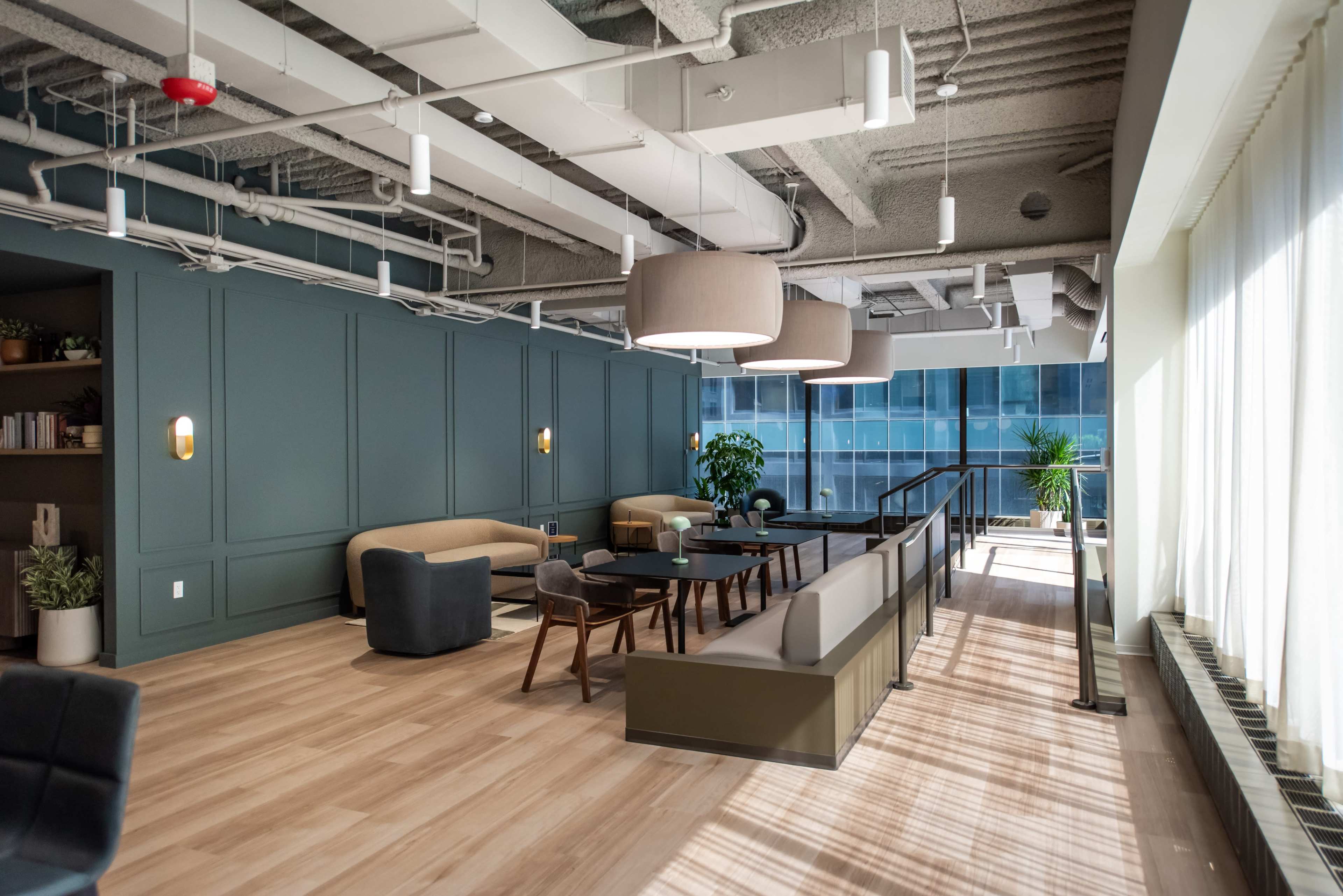 The image shows a modern office lounge area with seating, a large window, and contemporary lighting fixtures.