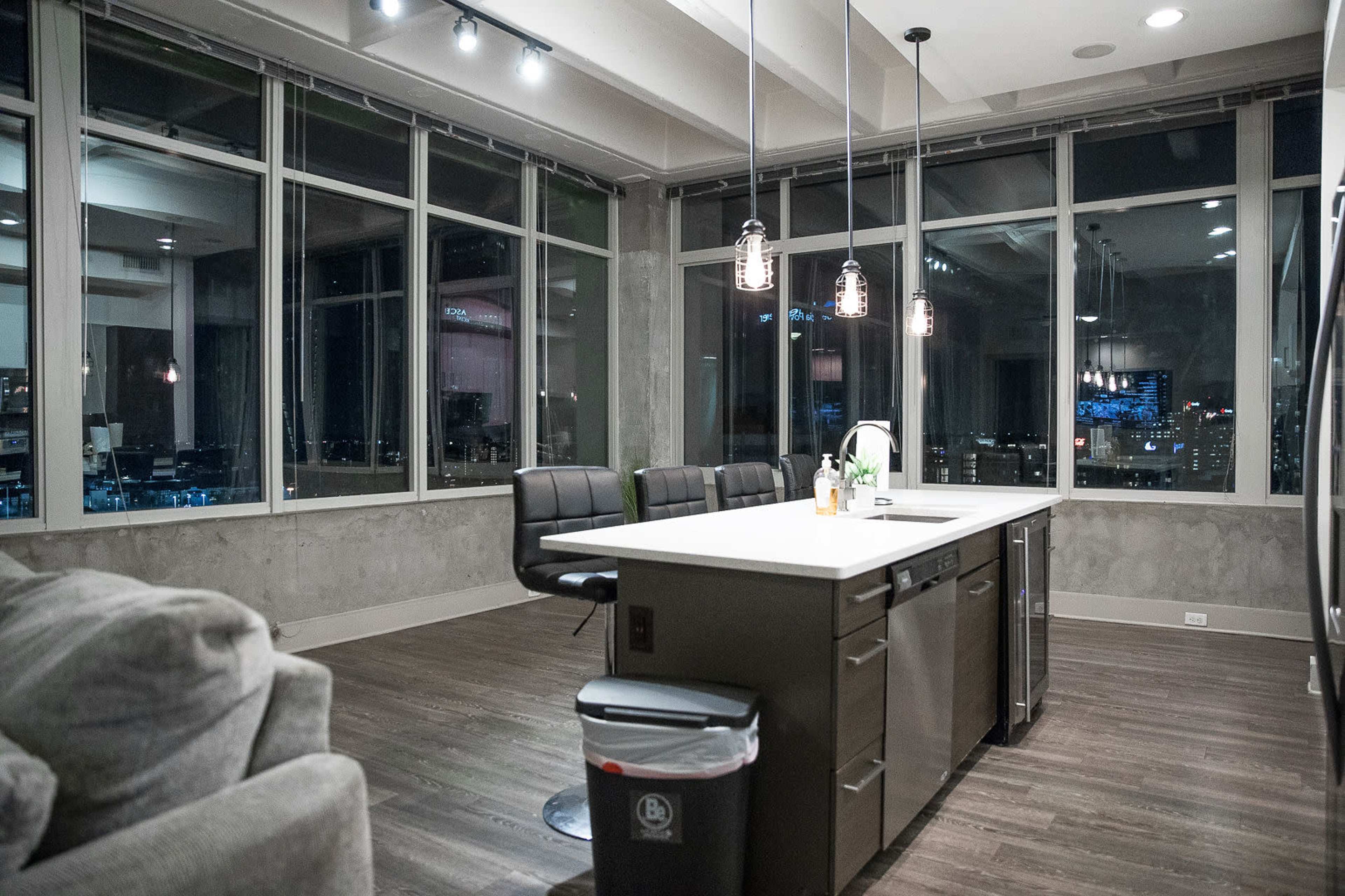 A modern kitchen with large windows offers a view of a city skyline, featuring an island counter and seating area.