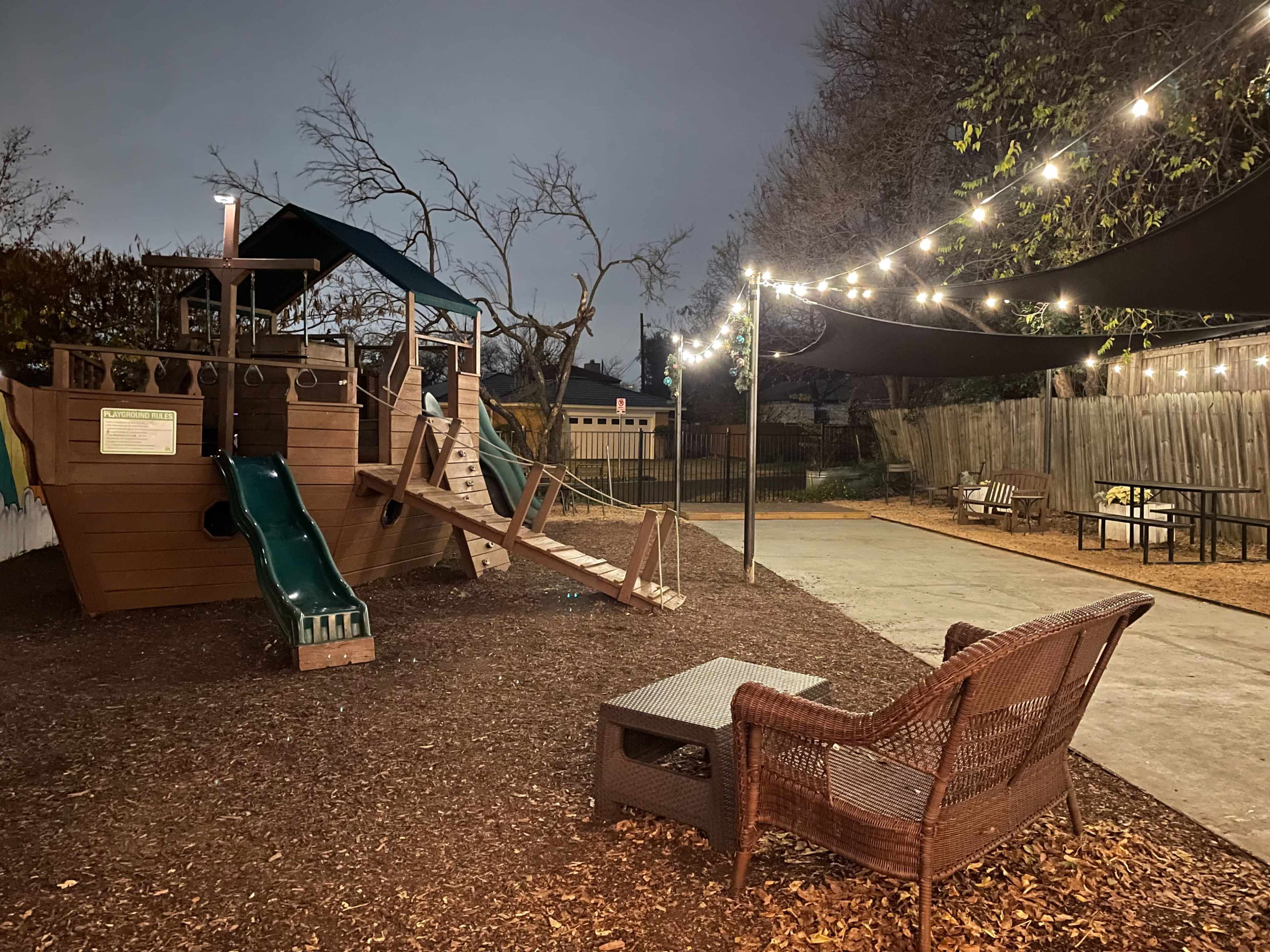 A playground with a slide and climbing structure is illuminated by string lights in a darkening outdoor setting.