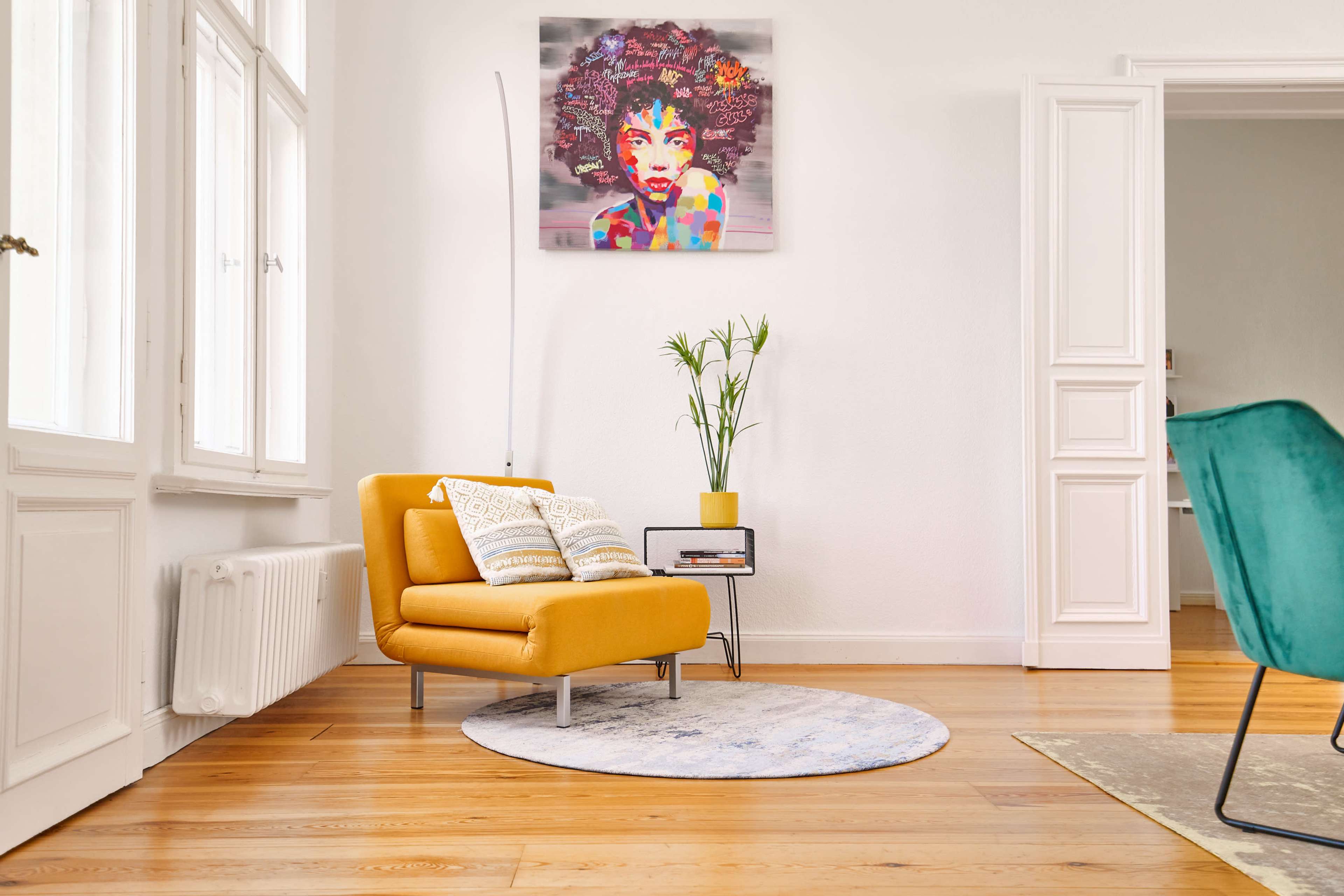 A yellow armchair with patterned pillows sits on a round rug in a brightly lit room with large windows and a colorful artwork on the wall.