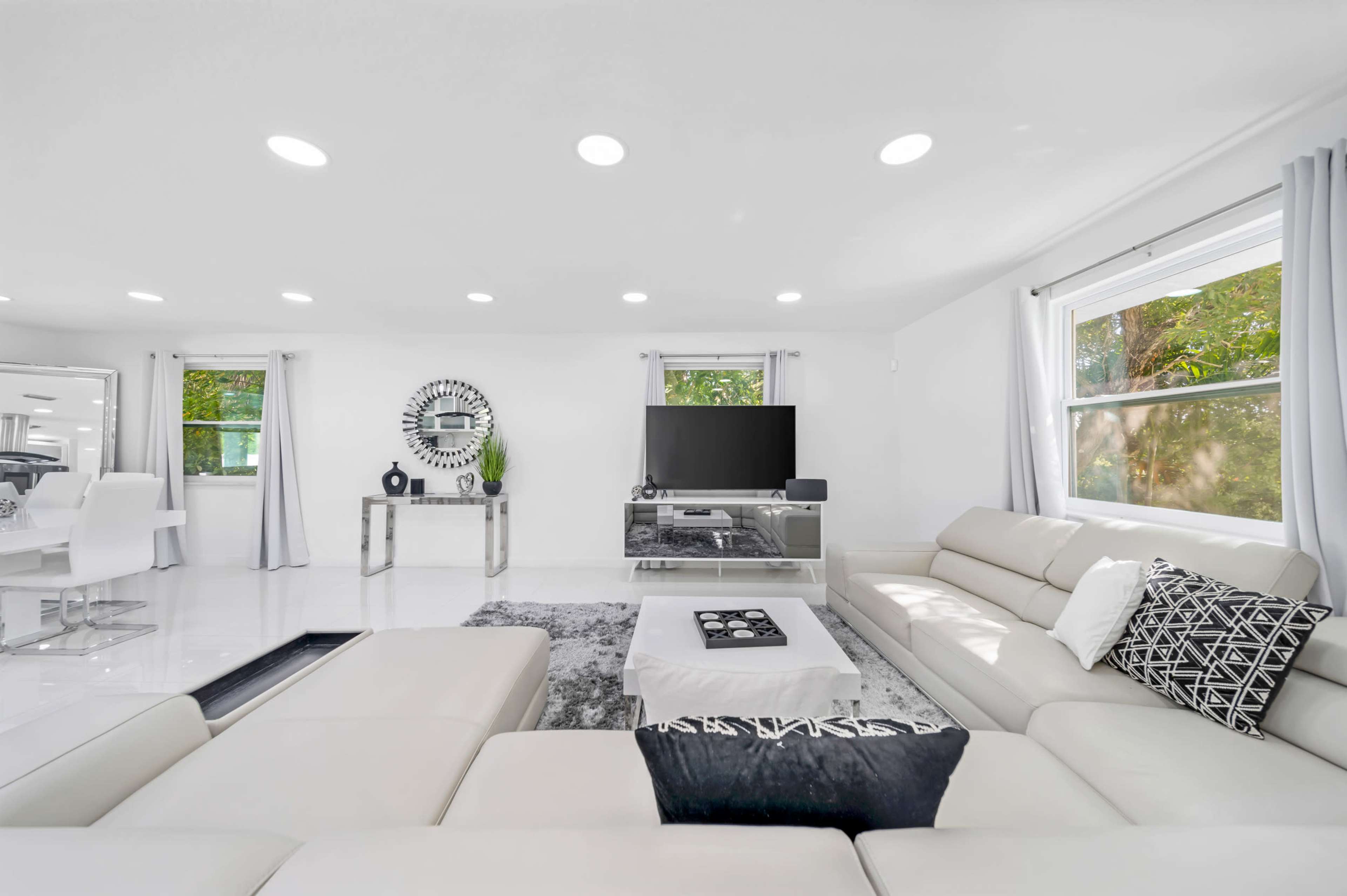 The image shows a modern living room with light-colored furniture, a large television, and a glass coffee table, surrounded by large windows allowing natural light in.