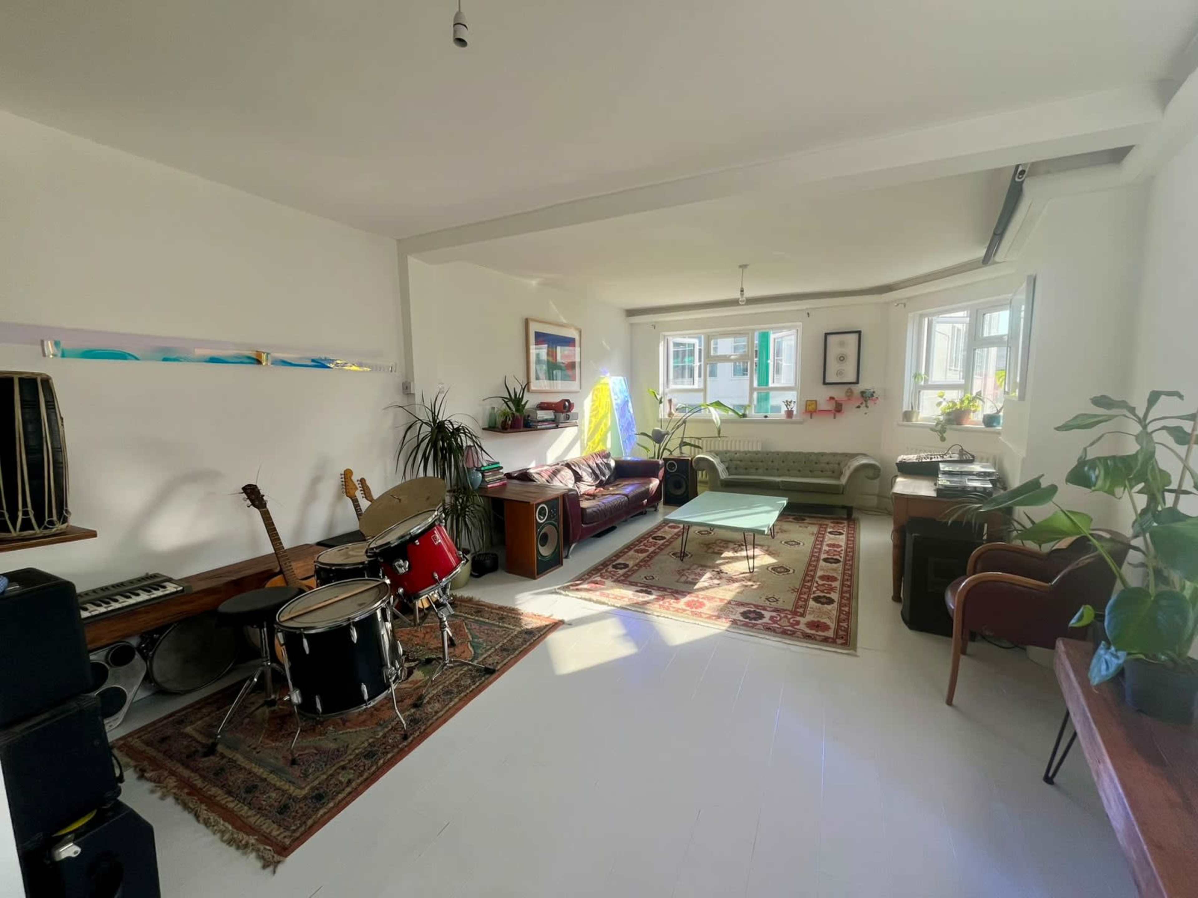 The image shows a bright, spacious living room with musical instruments, a patterned rug, plants, and large windows.