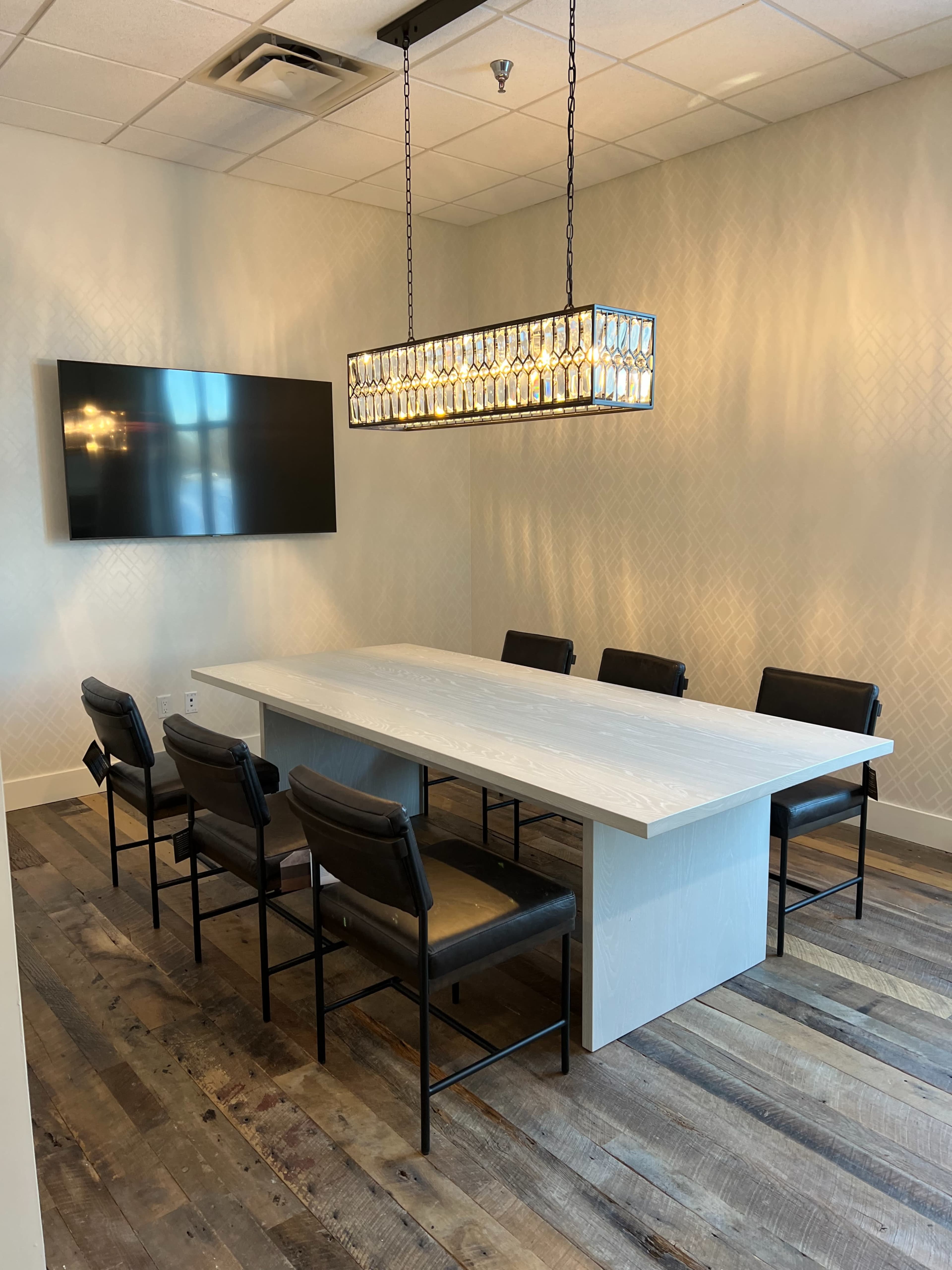 A modern conference room features a rectangular table surrounded by six black chairs, with a large wall-mounted screen and an elaborate light fixture overhead.