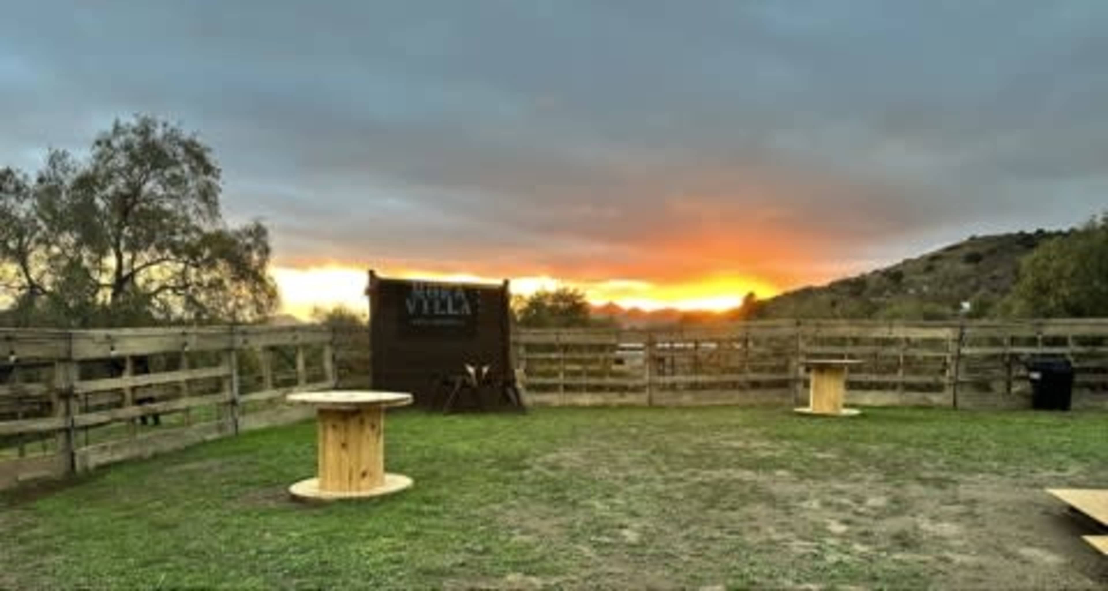 The image shows a rustic outdoor space with wooden tables and a fence, set against a backdrop of a sunset over hills.